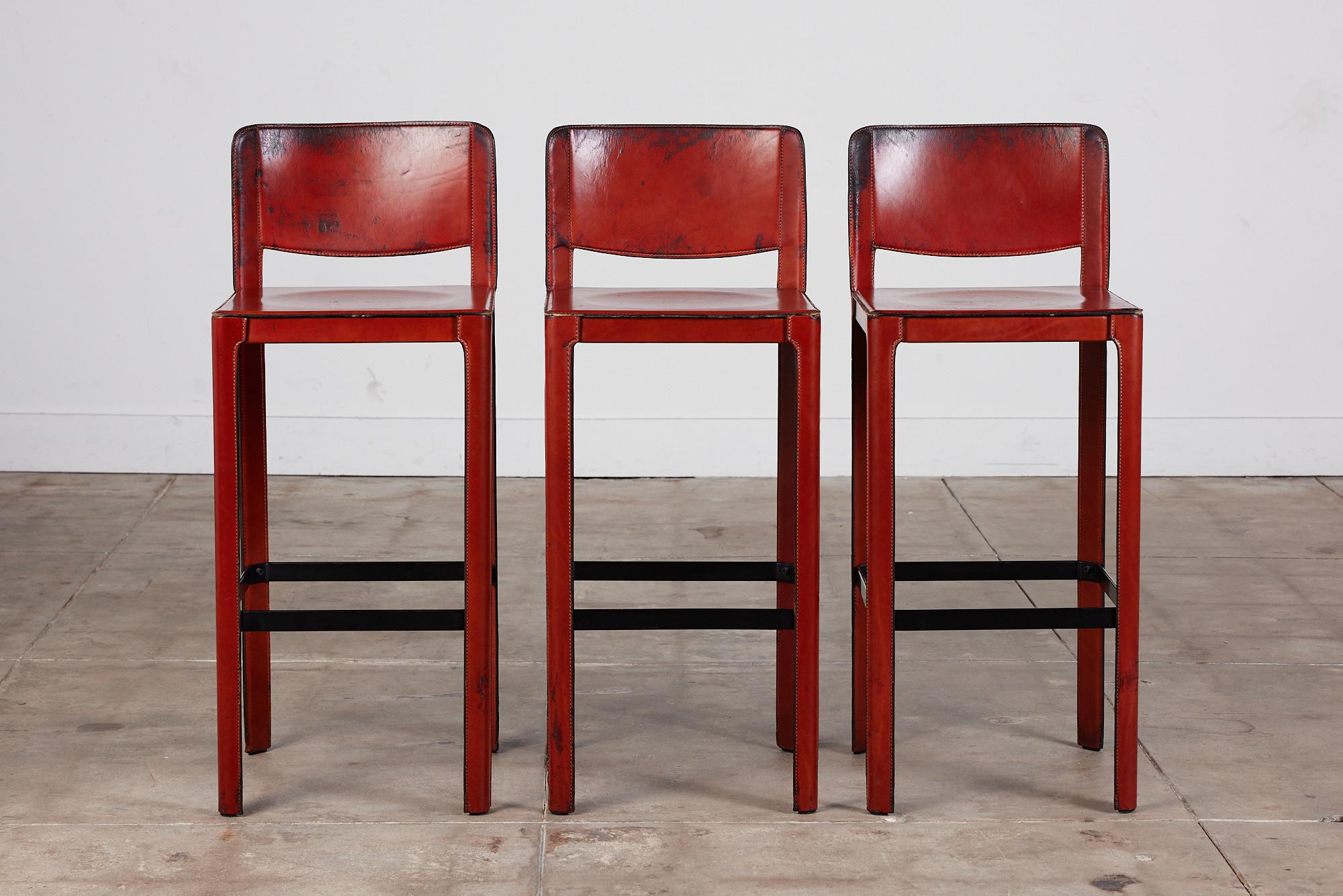 Matteo Grassi bar stool c.1980s, Italy, features a patinated reddish orange saddle leather which is wrapped atop a steel frame with stitched detailing. A black enameled square metal support bar connects all four legs and can be used as a footrest.