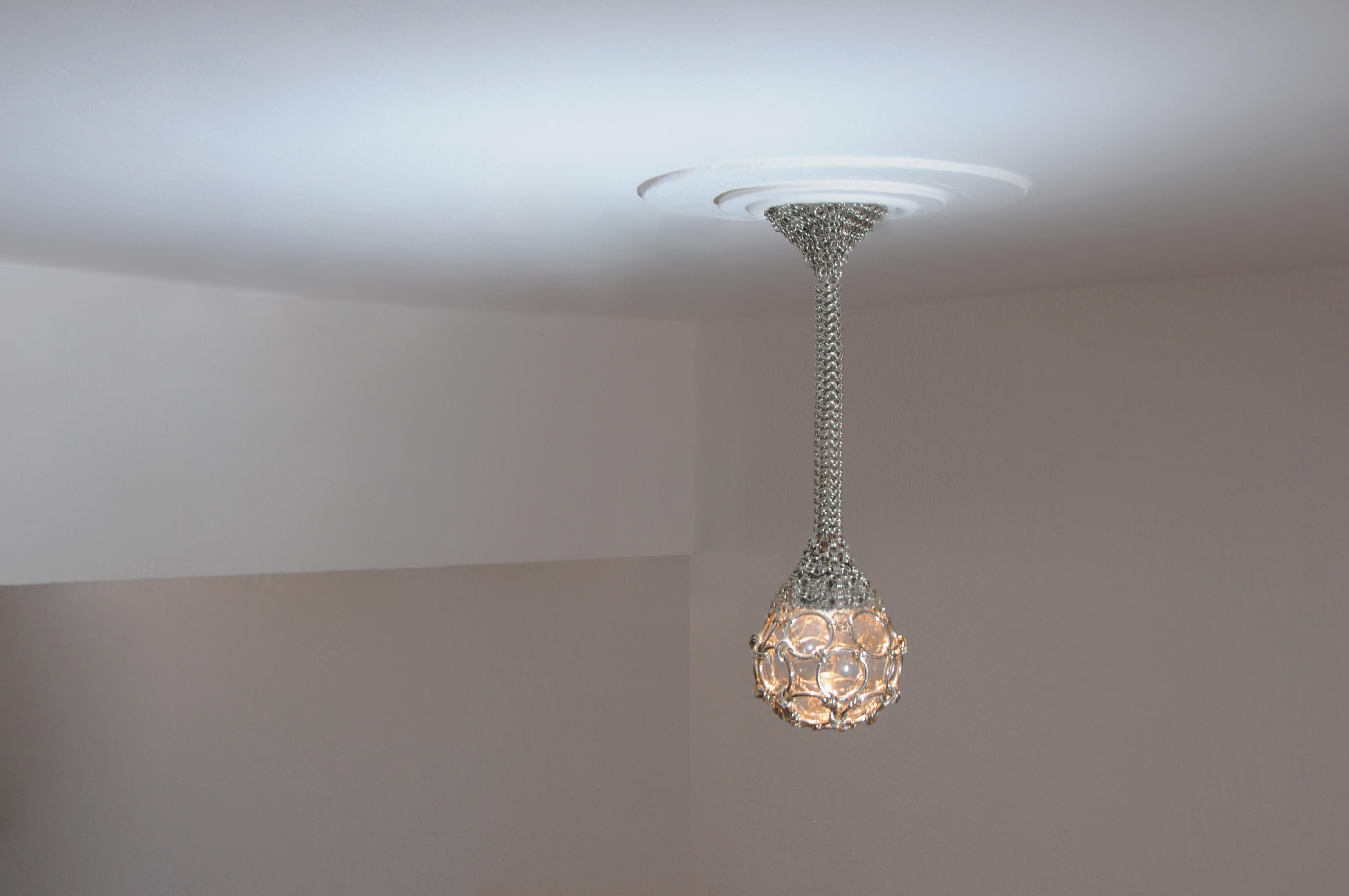 Single Pendant Chainmail Droplet Sculptural Chandelier is a beautiful hand made light piece. A chainmail web hugs the lamp’s canopy, as the knit gets tighter the chainmail envelopes into a rope which hangs to finally suspend a glass globe. All the