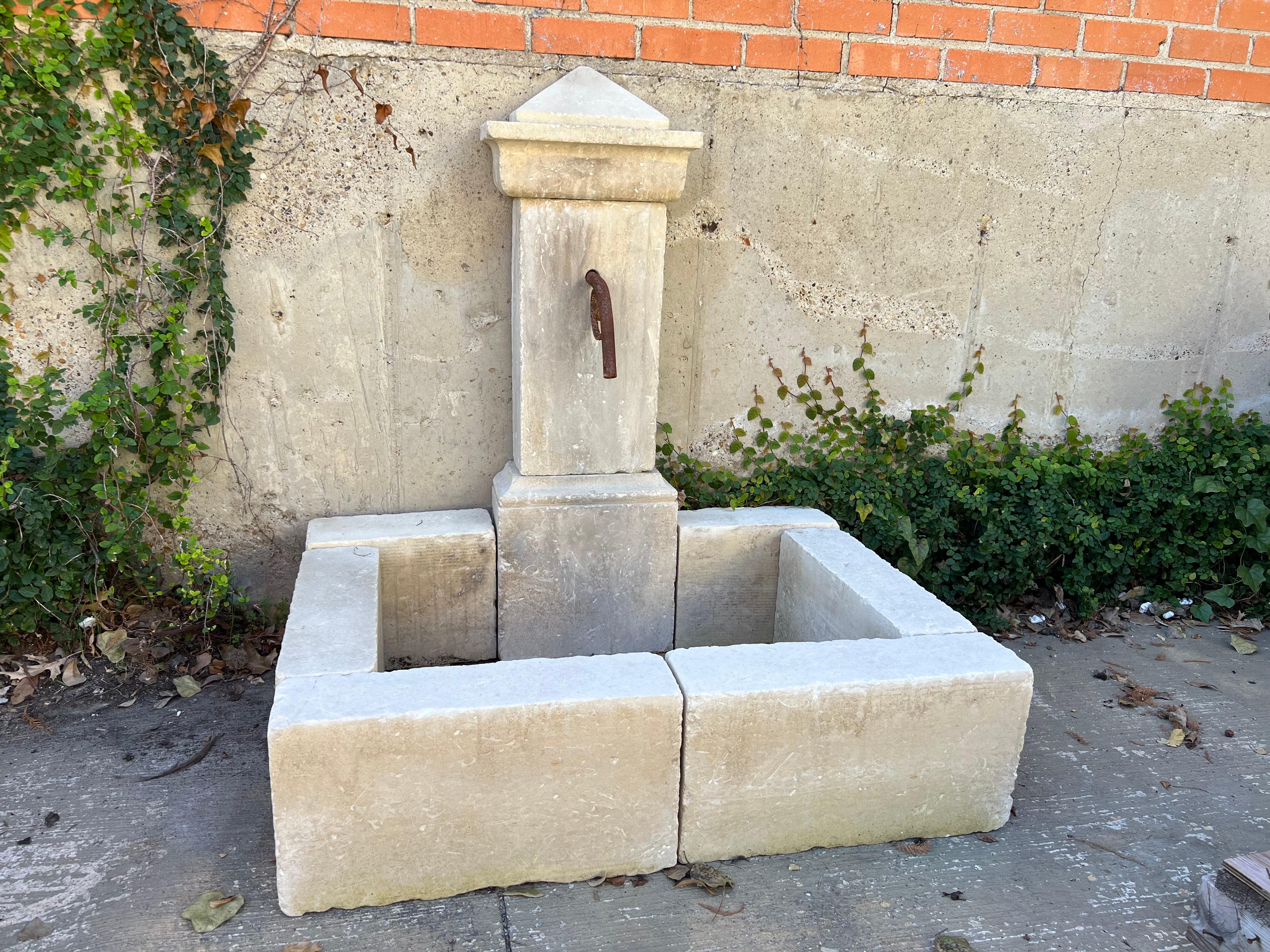 This hand-carved, single-pillar wall fountain consists of 10 pieces of southern Italian limestone. The fountain is topped by an angular pyramid finial that rests above an entablature with a deep cavetto cornice. An elaborately scrolled iron spout
