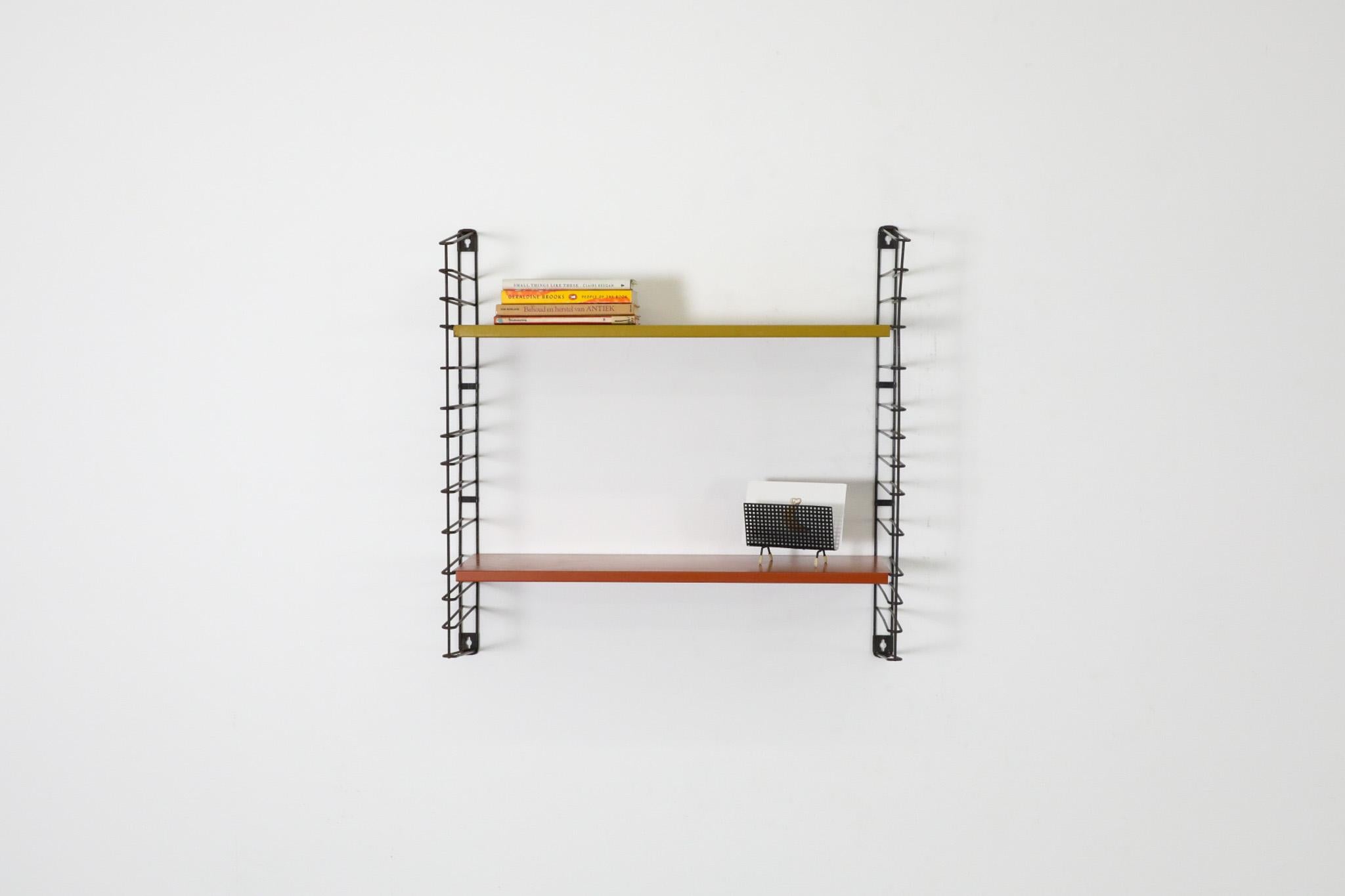 Estantería mural Tomado de una sola sección, con estantes de color naranja quemado y dorado sobre montantes de alambre de metal esmaltado en negro. En estado original, con desgaste visible propio de la edad y el uso. Hay otras estanterías similares