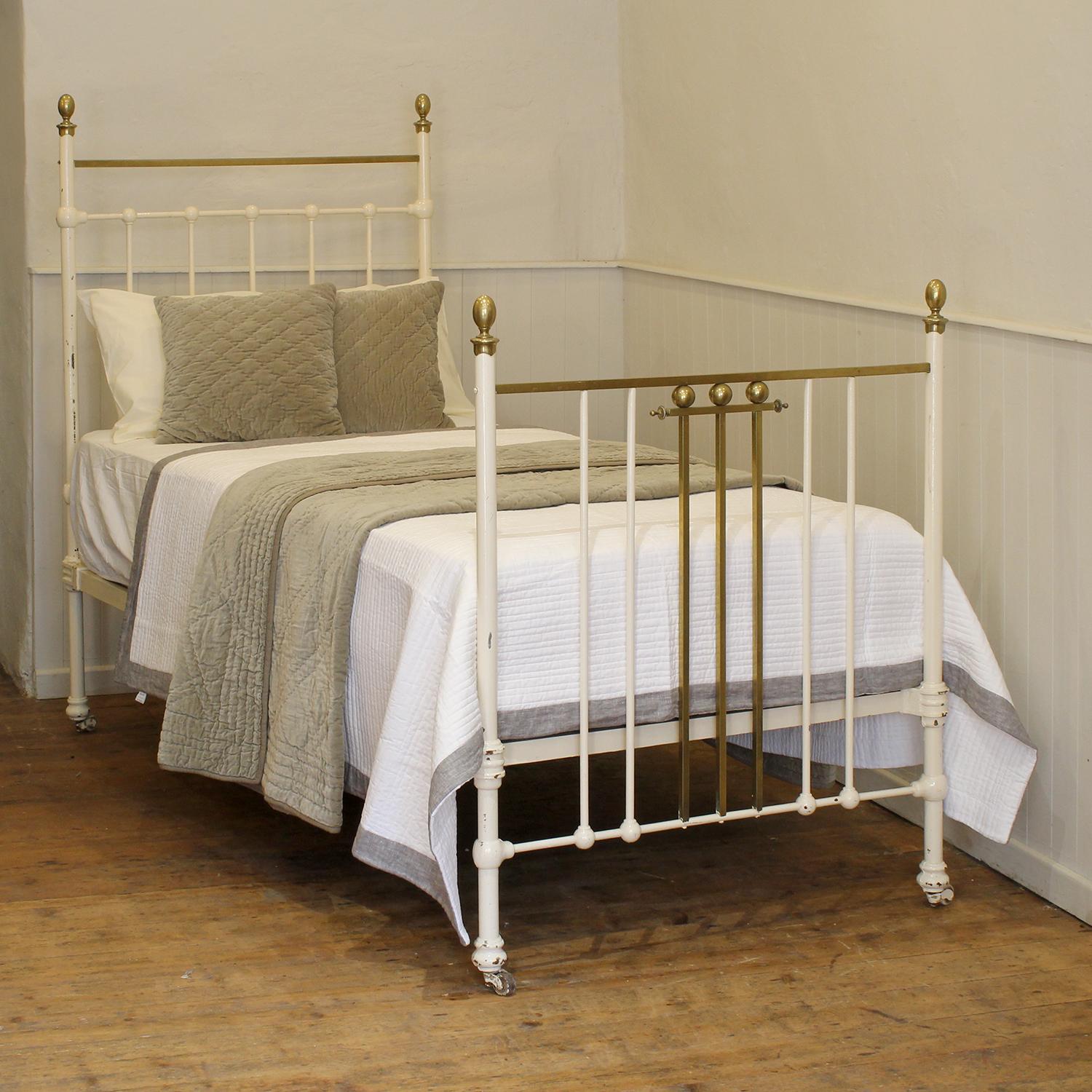 A traditional style Edwardian antique bed painted in white, with square section brass top rail, decorative features in the footboard, and oval brass knobs. 
The bed shows signs of its age with some marcks in the paintwork. We can leave these for