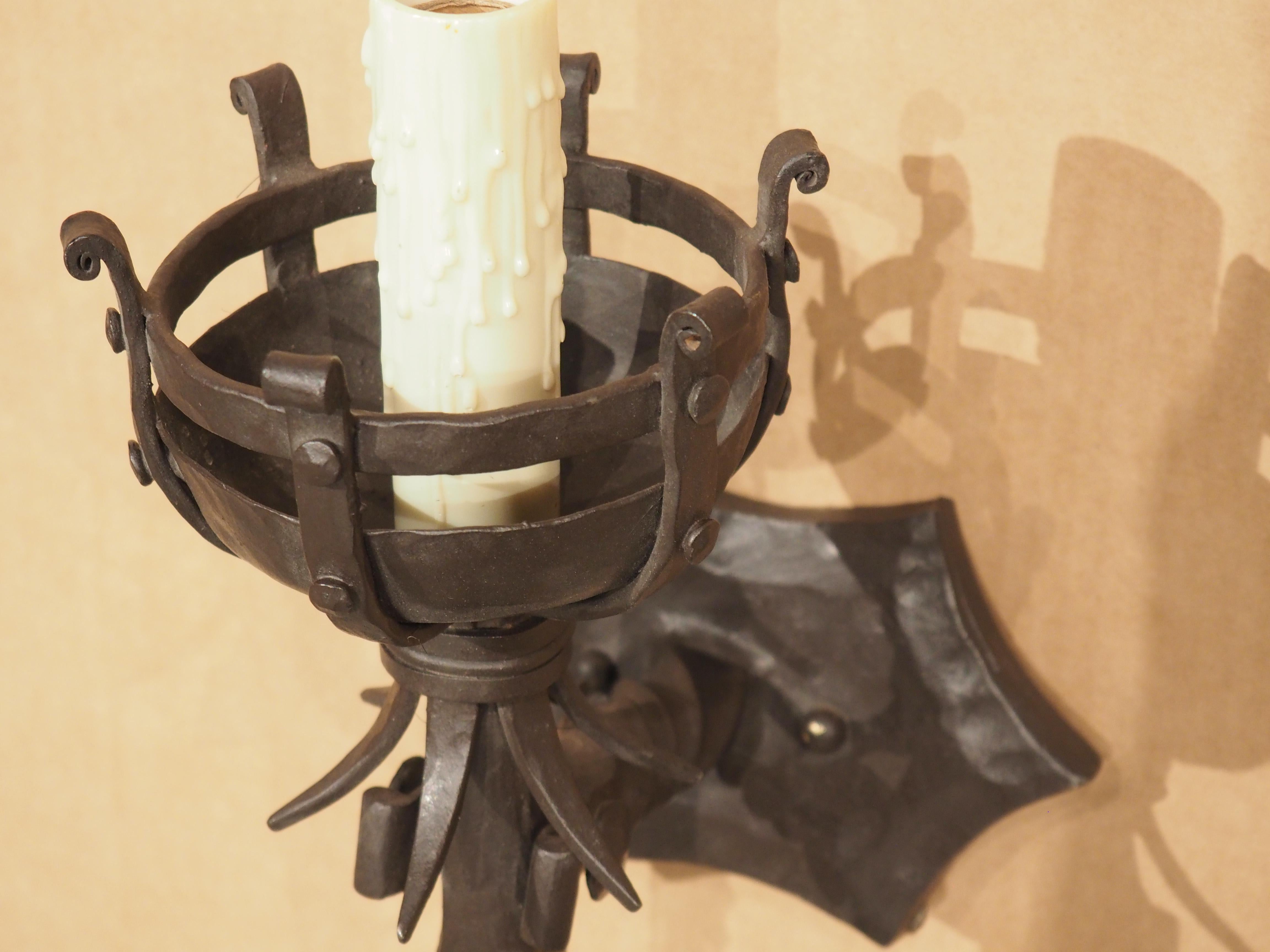 Resembling a Medieval-style castle torch, this single wrought iron wall sconce blends functionality with a unique aesthetic appeal. An expansive saucer bobeche encompassed within a basket-like cup comprised of scrolled iron supports houses an