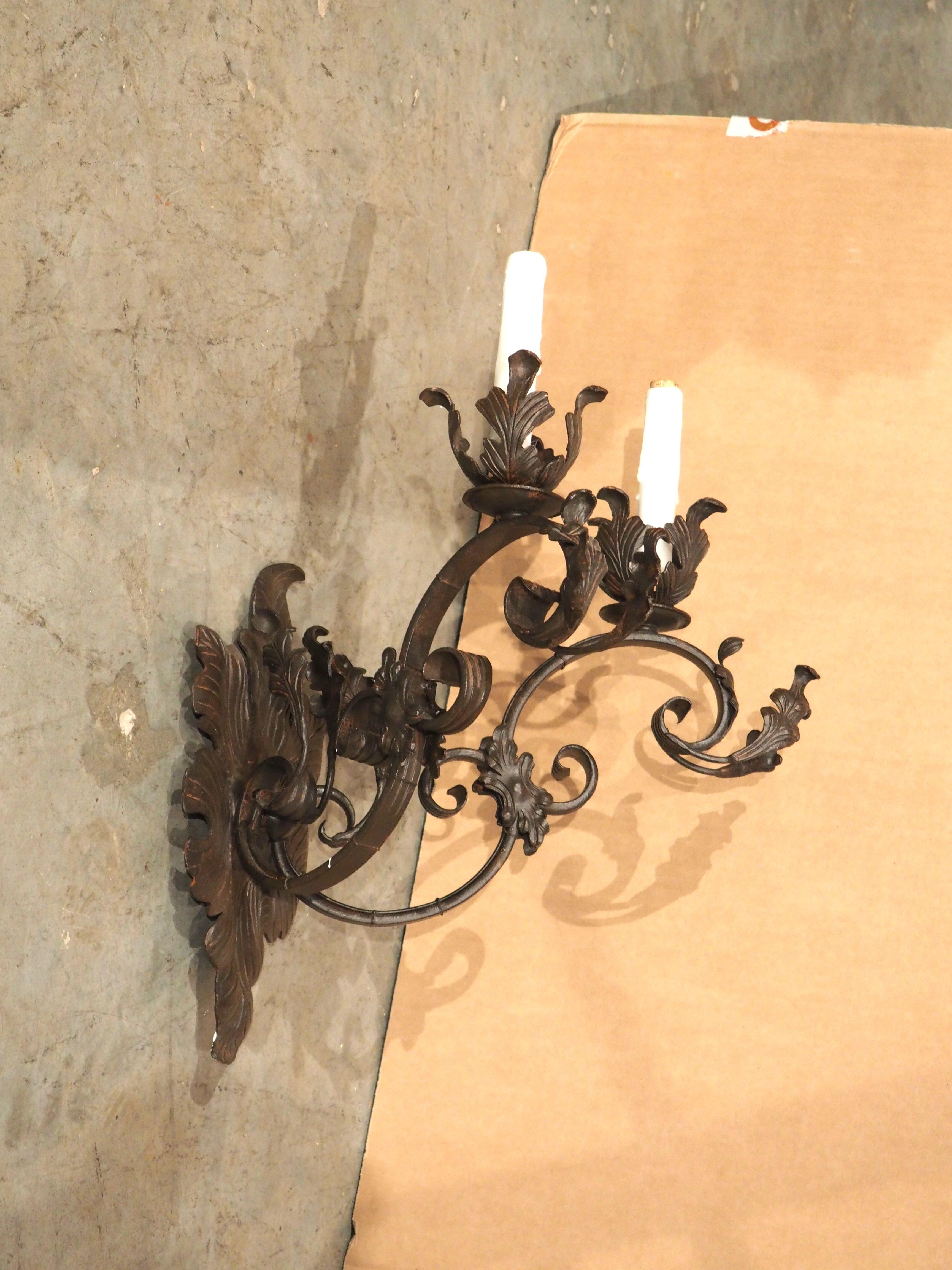 An elaborate display of foliate ornamentation adds to the beauty of this wrought iron wall sconce. Each of the two white faux candle sleeves is partially enveloped by a parted leaf bobeche above an S-scroll. The highly sinuous arms have additional
