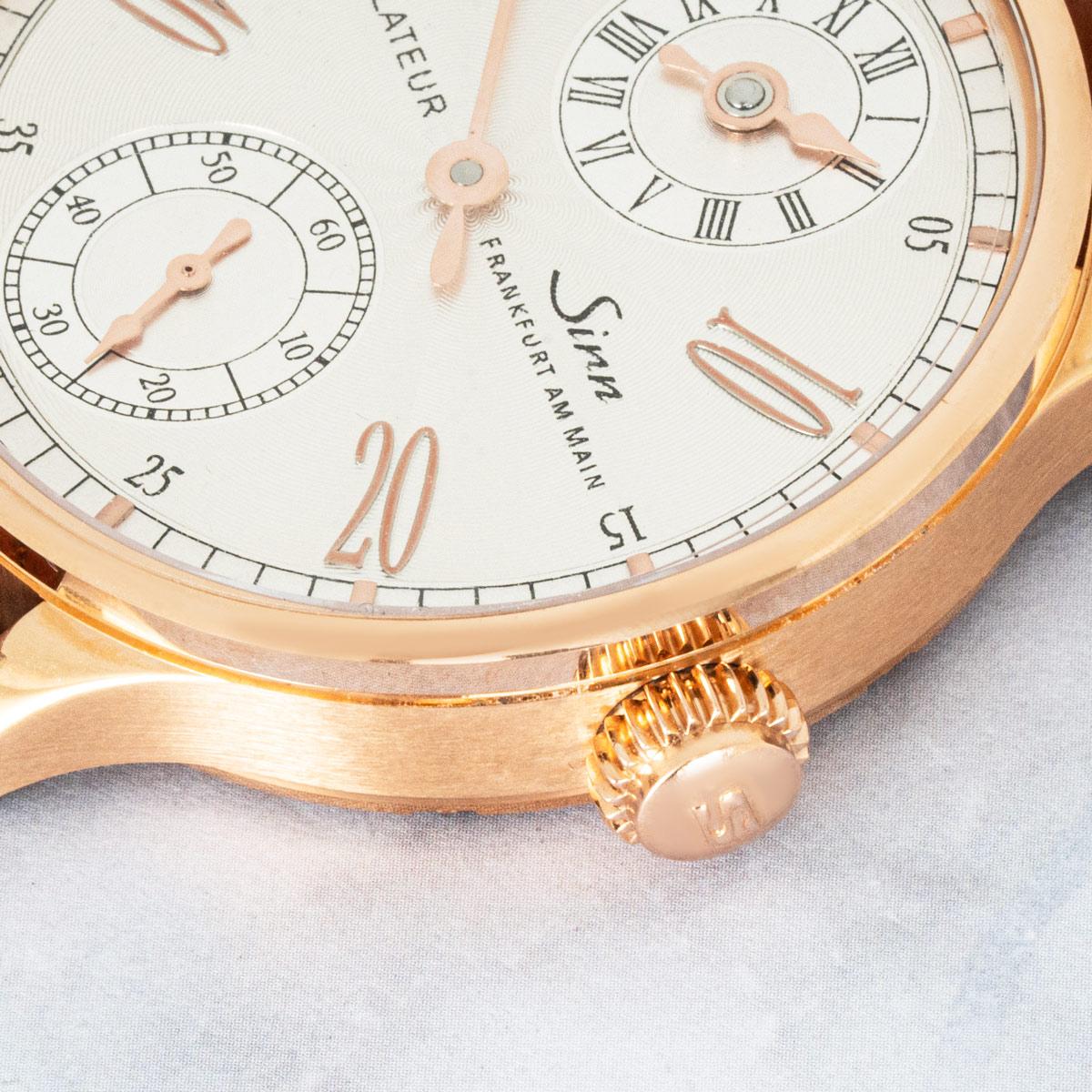 A rose gold 44mm Regulateur wristwatch by Sinn. Features a silver guilloche dial with the hour sub-dial at 12 o'clock, central minutes, and a small seconds sub-dial at 6 o'clock.

The watch is presented on a brown leather Sinn strap (additional
