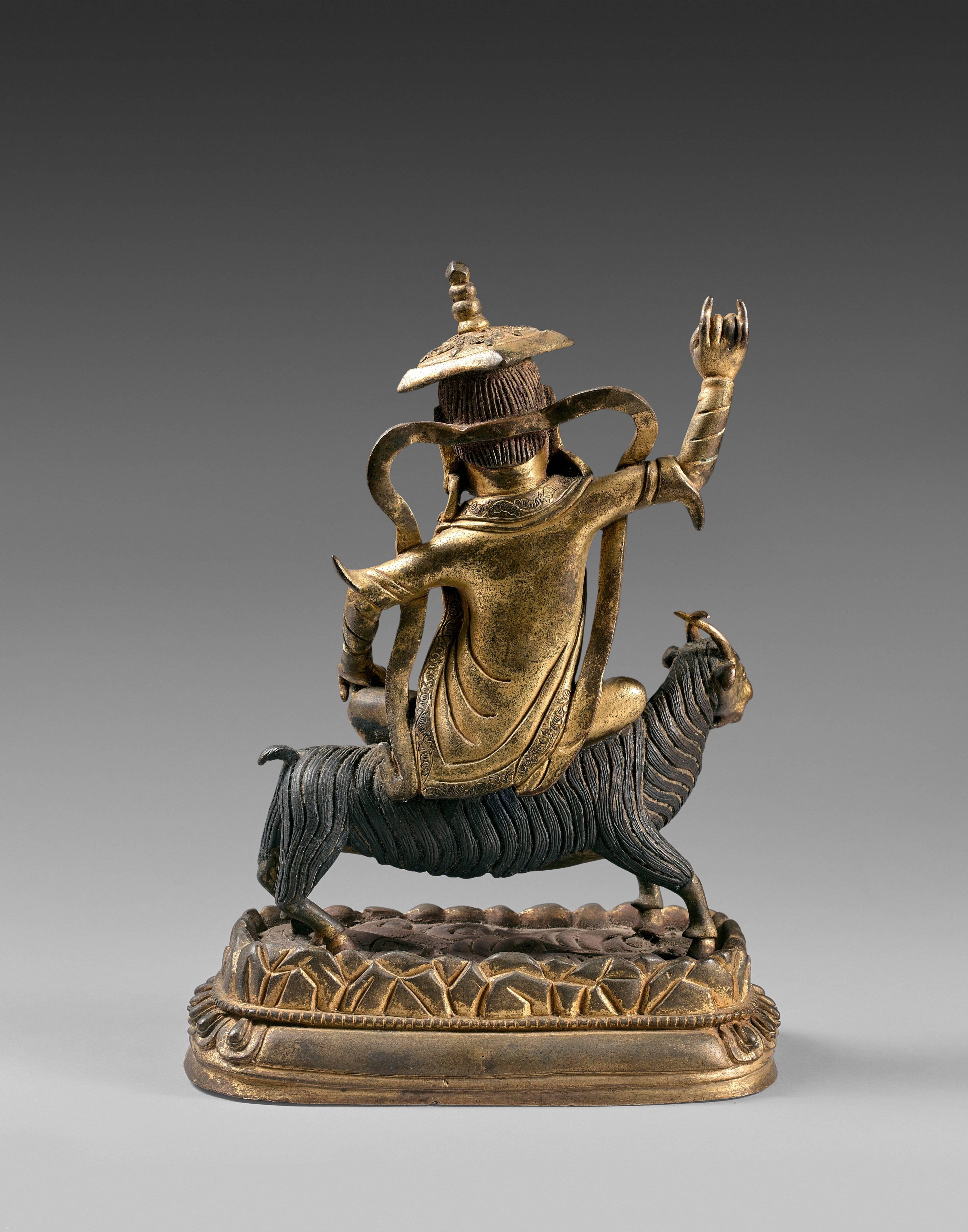 The wrathful divinity Dam-Can ridding a goat, dressed with a traditional Tibetan robe and a square hat, with a fluttering scarf, the ferocious face with an open mouth showing fangs. The divinity and the animal on a rocky base and a lotus pedestal.