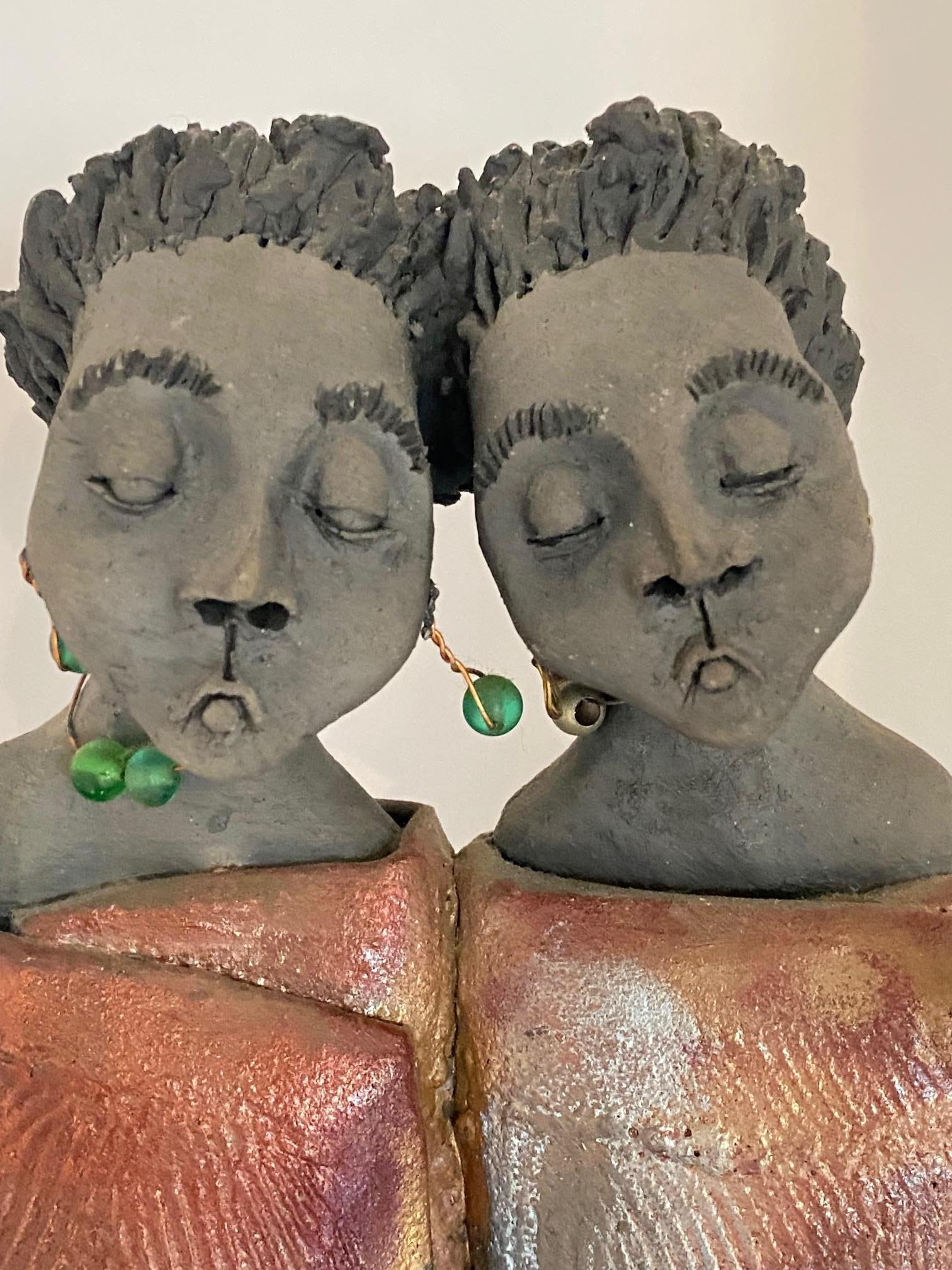 This sculpture is a beautiful ceramic piece depicting two women leaning affectionately toward each other. The women are wrapped in a large, unique cloak covering their bodies. The two women are wearing green earrings and have short hair. The