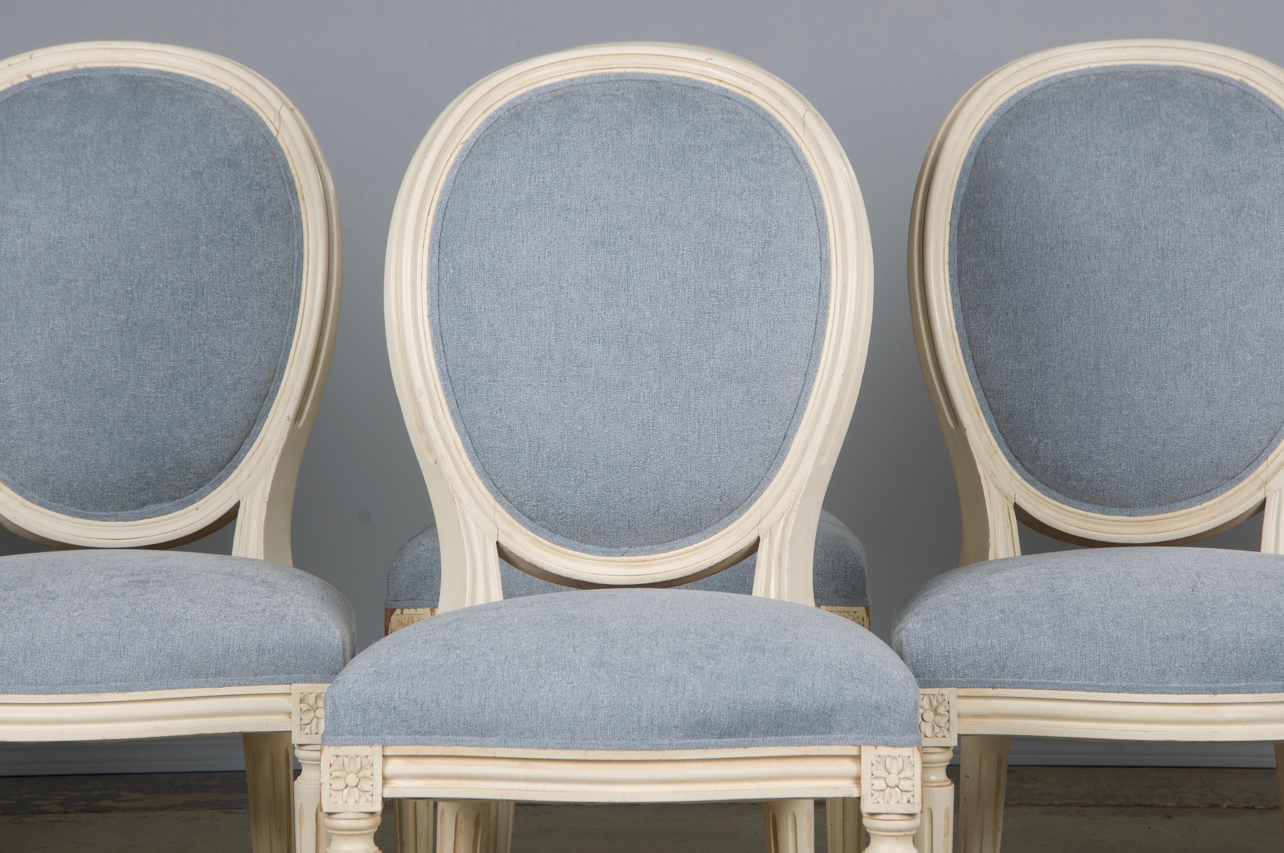 An exquisite set of six antique French dining chairs in the Louis XVI style, sourced directly from Provence, France. Each chair features a hand-carved, oval-shaped medallion backrest finished in creamy antique white paint with a naturally aged