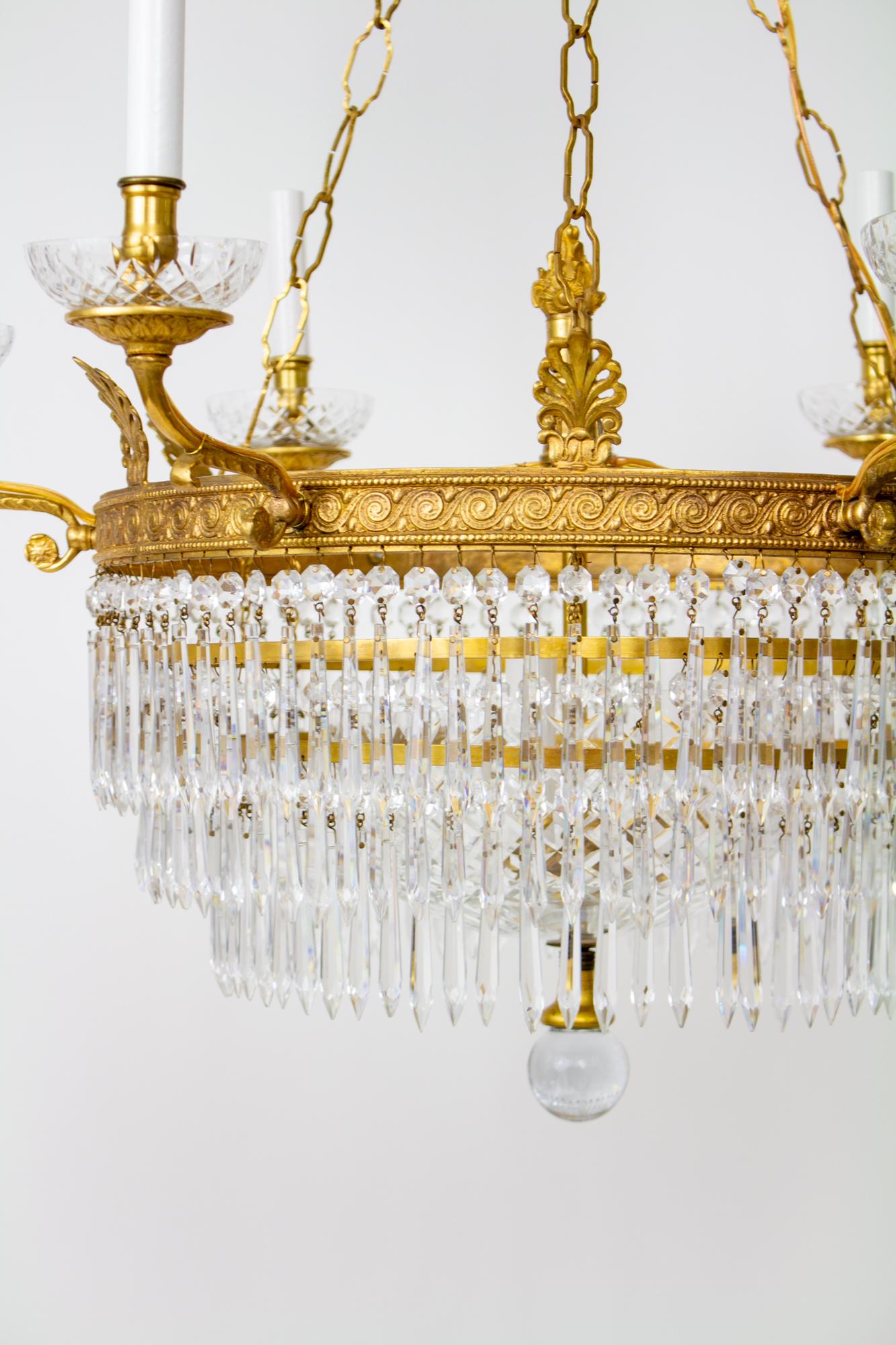 Six arm brass and crystal empire style chandelier. Gilt brass with Crystals. Central brass ring with classical wave cast design. Six arms extend from the central ring with cast design, cut crystal bobeches and candelabra sockets with candlecovers.