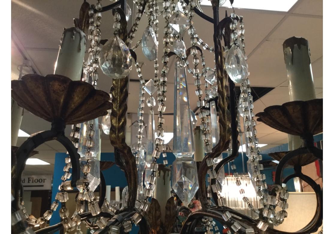 Questo lampadario si presenta con sei bracci a volute foliate, bobeches scanalate e smerlate, ghirlande di perline di cristallo, grandi prismi a lancia e cristalli a goccia drappeggiati da tre livelli e terminanti in un grande cristallo a goccia
