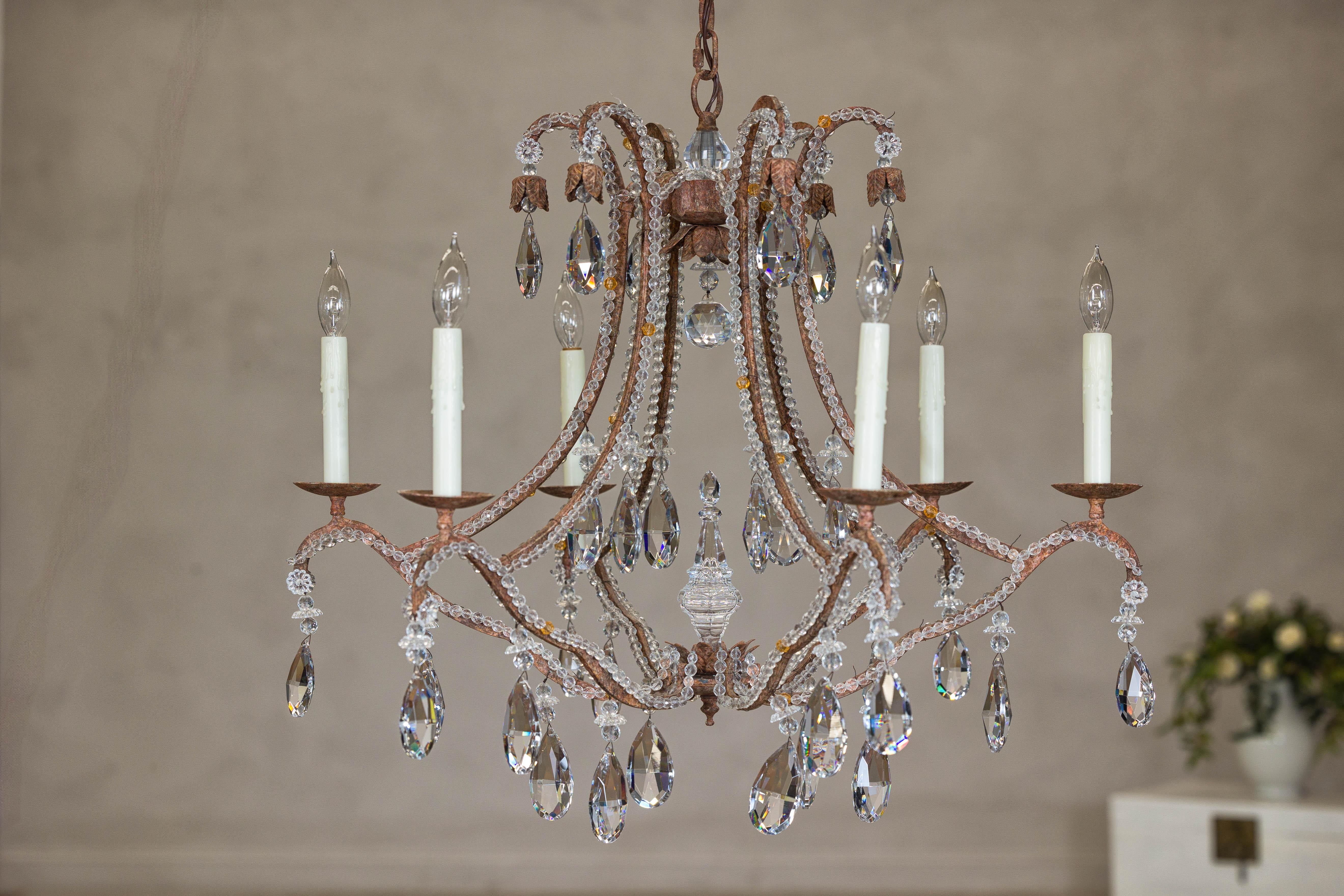 Dieser sechsflammige Kristallkronleuchter mit einer Struktur aus patiniertem Eisen und geschwungenen Armen verleiht jedem Raum einen Hauch von Vintage-Charme und Eleganz. Die anmutigen Arme des Kronleuchters, die aus patiniertem Eisen gefertigt