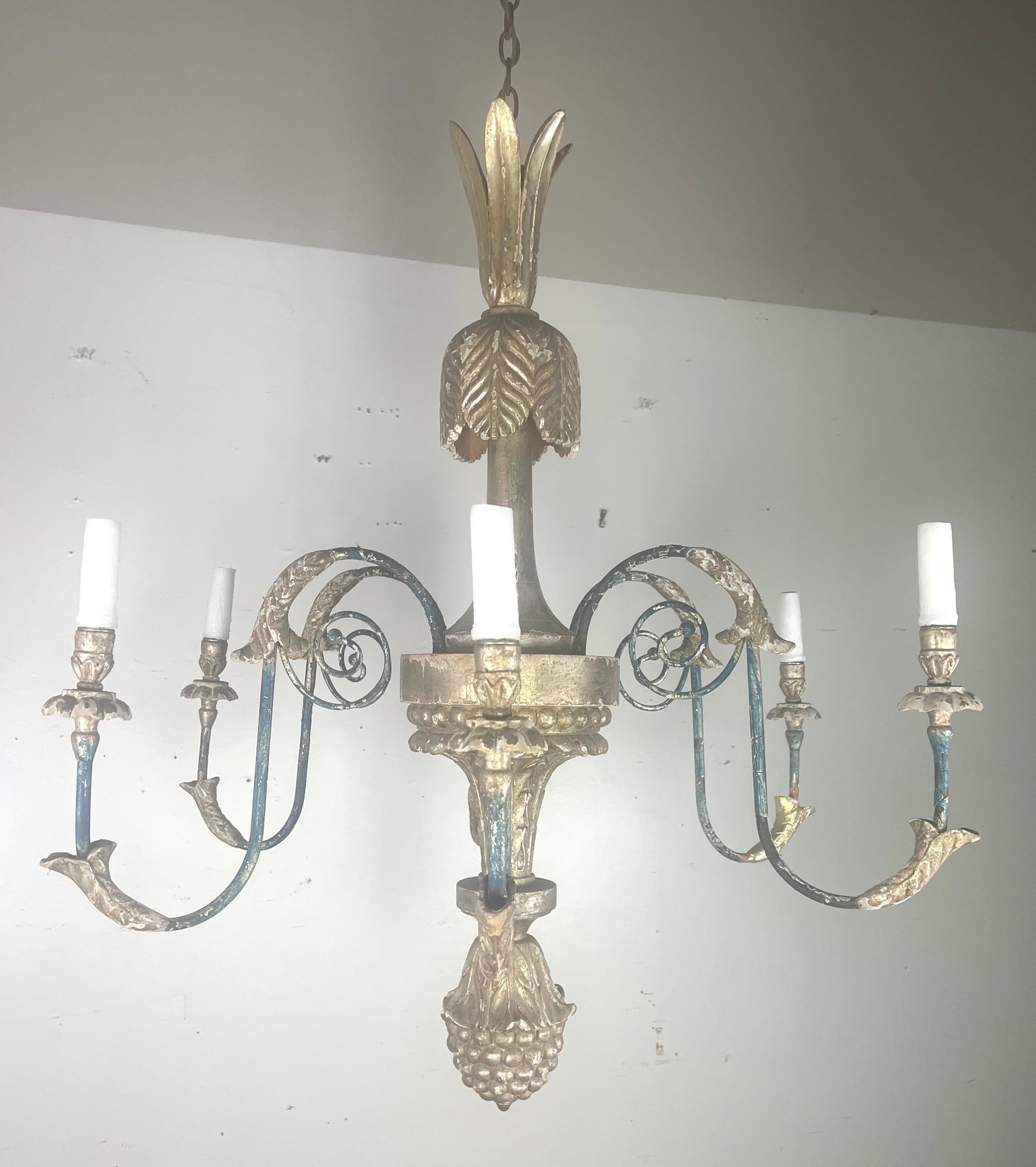 Questo lampadario tradizionale in stile italiano presenta una colonna centrale ornata da un terminale superiore a forma di ananas, che aggiunge un tocco di unicità al suo design.  Il lampadario ha sei bracci metallici curvi, ognuno dei quali