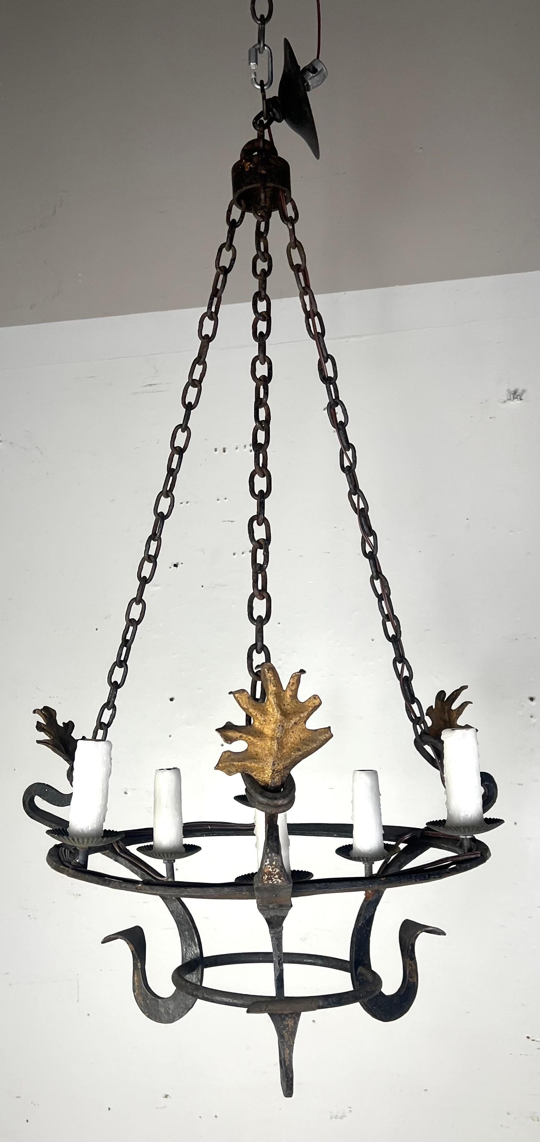Si tratta di un lampadario rustico in ferro battuto con un design di ispirazione medievale.  Presenta una struttura circolare sostenuta da quattro catene che convergono in alto per essere appese.  Il lampadario è dotato di sei luci a candela con