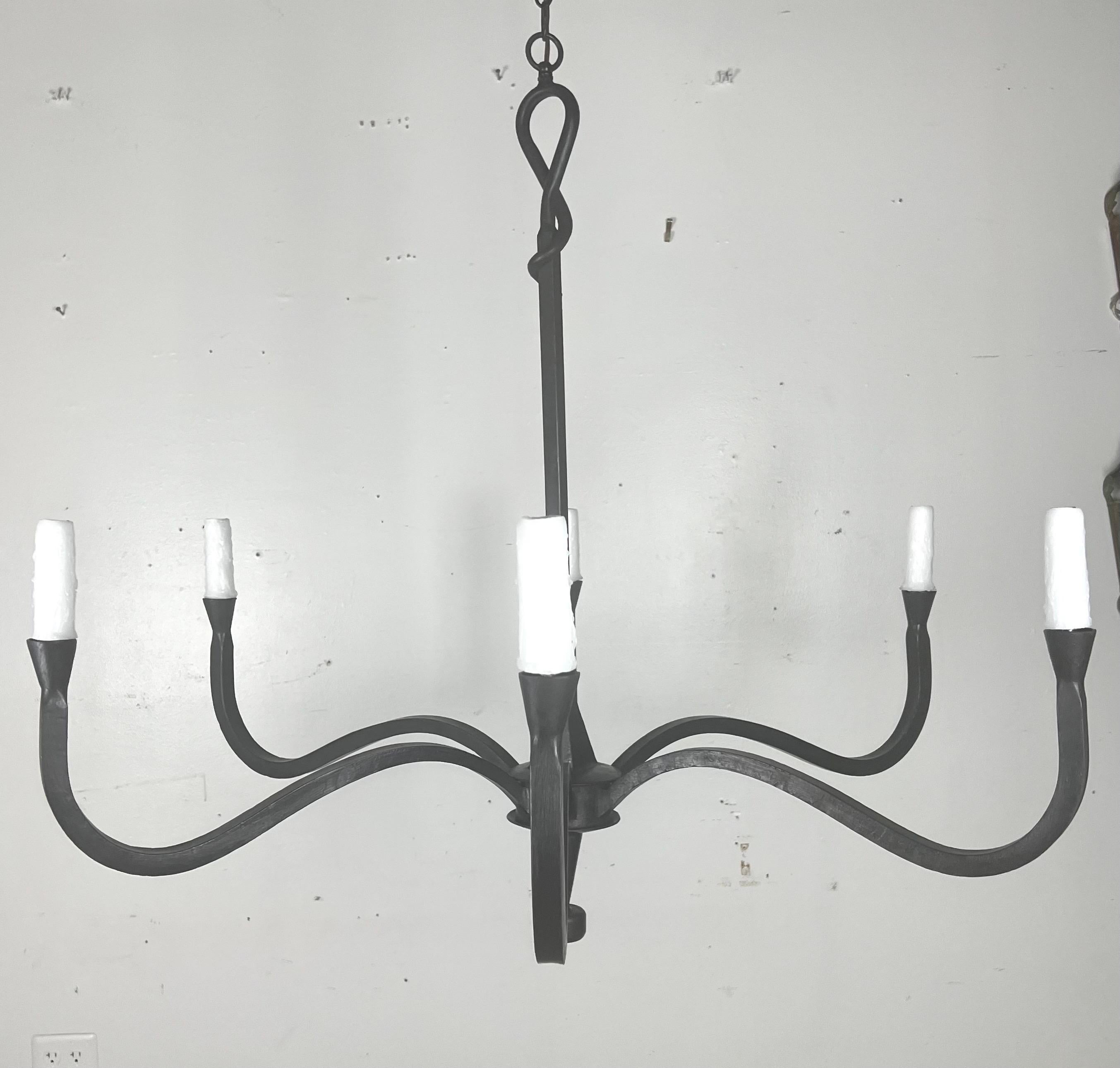 This six light chandelier has a minimalist and elegant design, crafted from wrought iron. It features six arms, extending symmetrically from a center hub, each ending in a drip wax candle cover.  The smooth curves and simple lines contribute to its