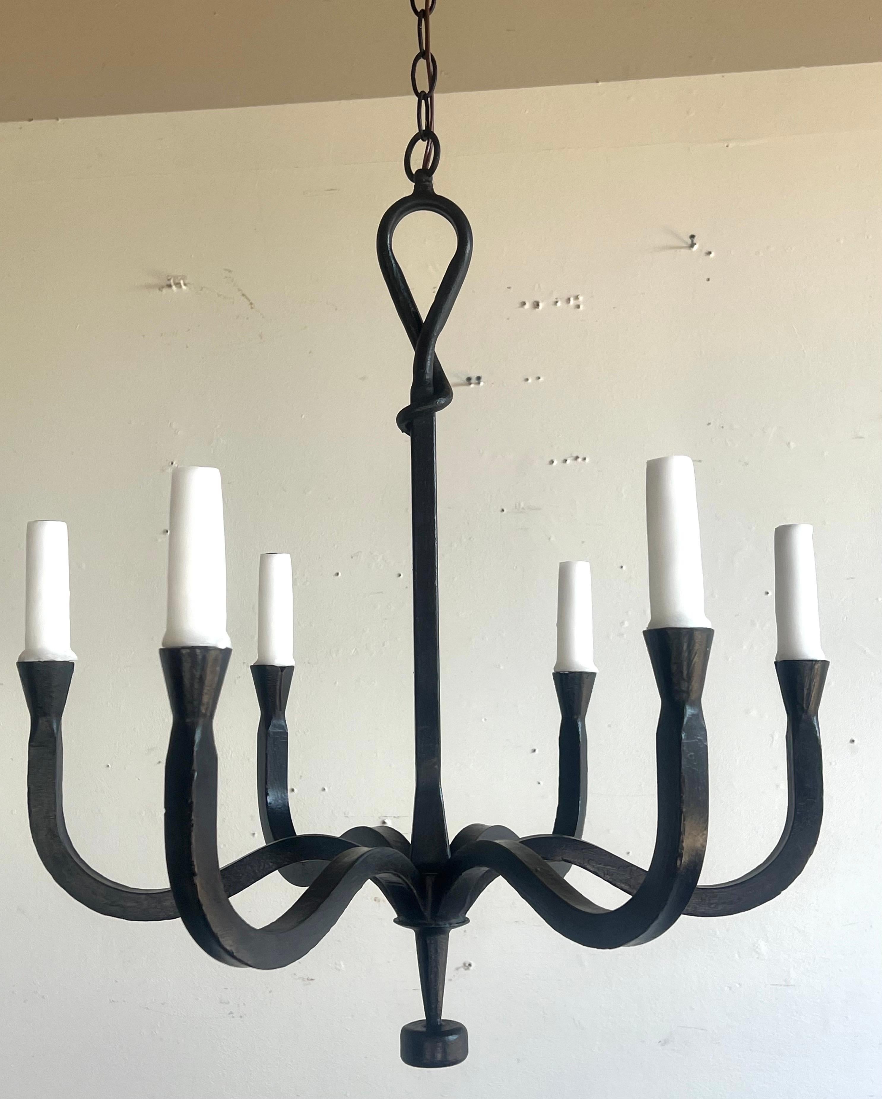 This six light chandelier has a minimalist and elegant design, crafted from wrought iron. It features six arms, extending symmetrically from a center hub, each ending in a drip wax candle cover.  The smooth curves and simple lines contribute to its