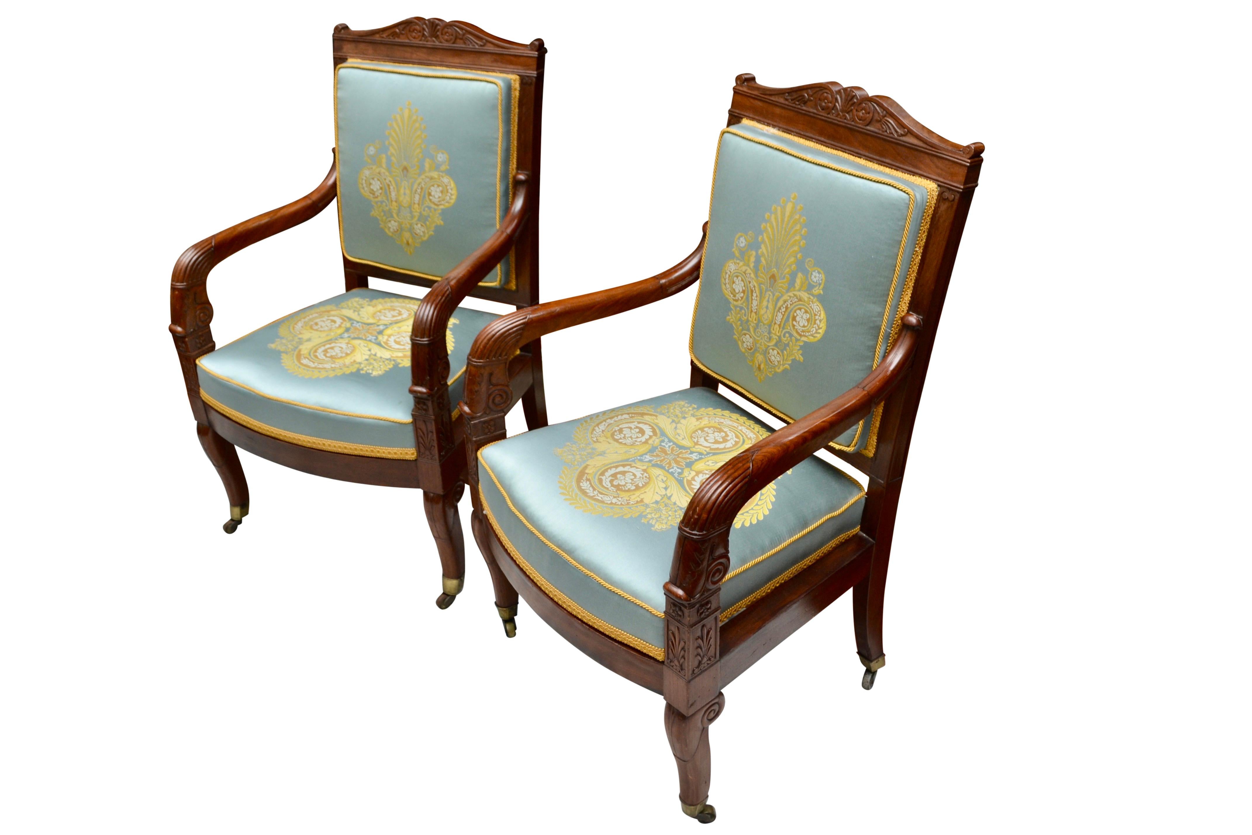 A classic pair of French Empire period open armchairs in well figured and carved mahogany; each with a square back topped by a bow shaped carved frieze featuring folliate rosettes and downscrolled arms. Where the arms join the chair rail there are