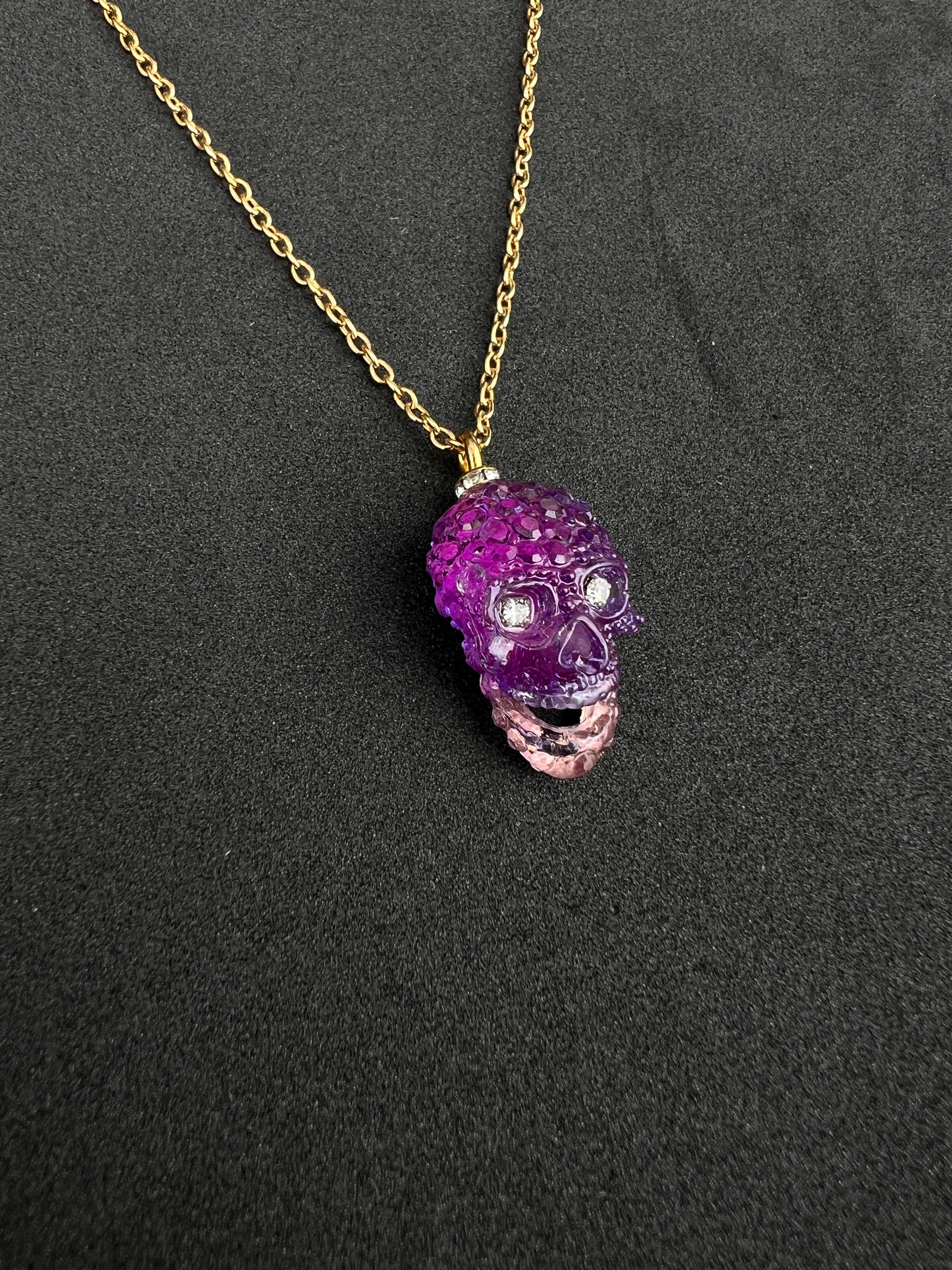 Questa splendida collana con ciondolo è dotata di una catena d'oro che non si appanna e che garantisce una lucentezza e una durata durature. Sospeso alla catena c'è un teschio in resina viola, intricato e impreziosito da occhi in cristallo