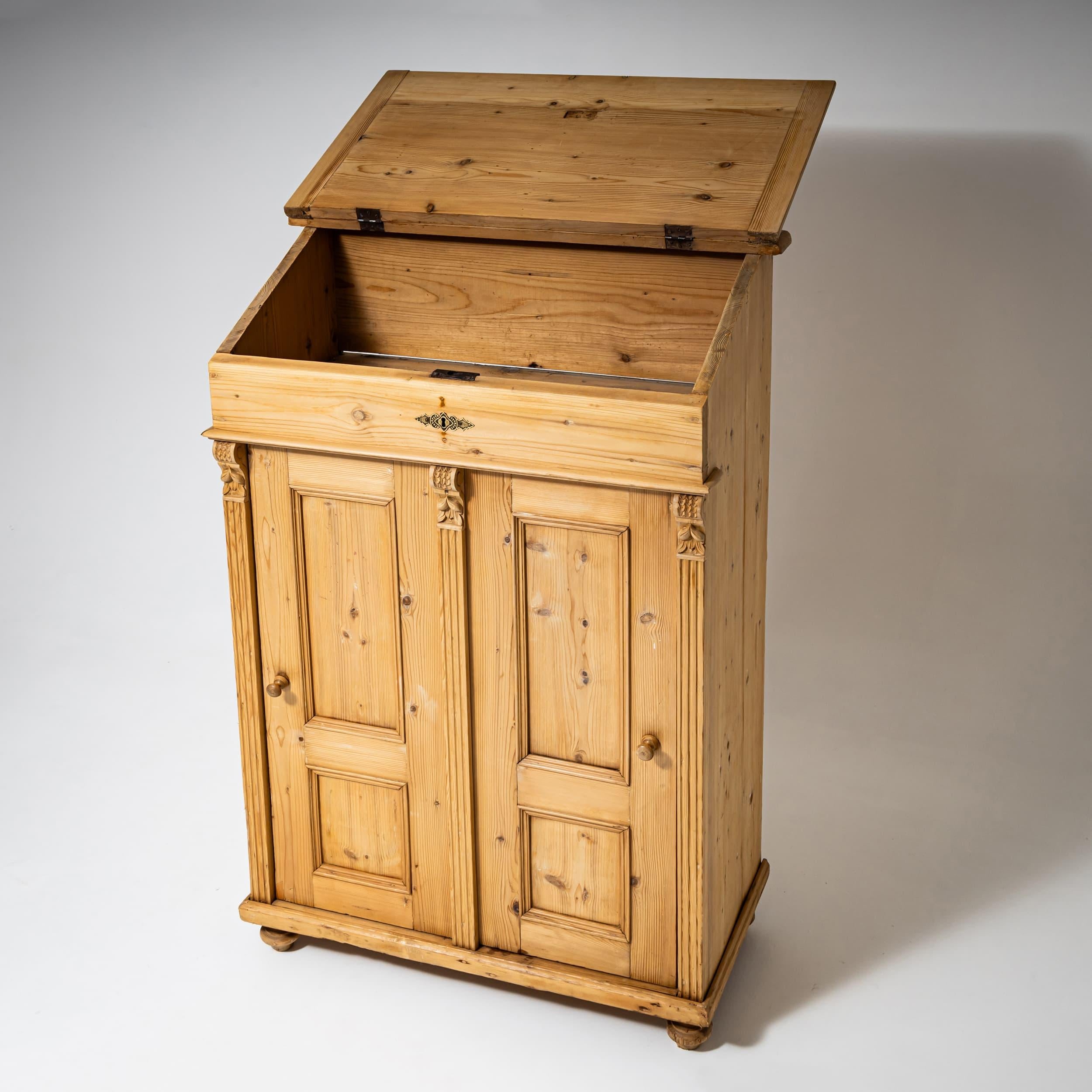 Slant-front desk in softwood with an angled writing flap revealing a spacious storage compartment beneath. Below, a two-door cabinet section features paneled doors with double fielding, flanked by fluted pilasters. The pilasters terminate in