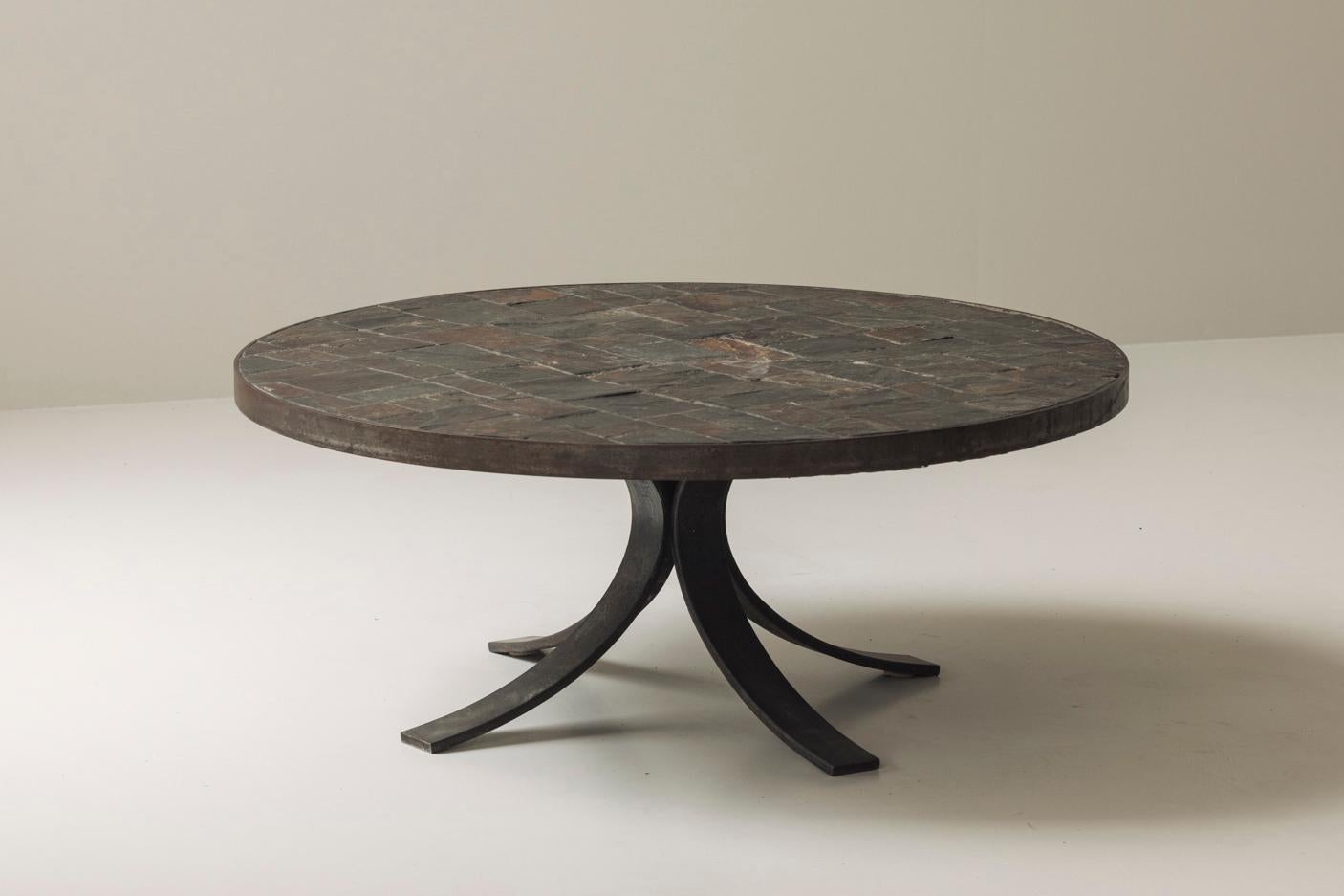 Cette élégante table basse française des années 1950 allie une matérialité brute à un design raffiné. Le plateau circulaire, fabriqué en pierre d'ardoise dans une incrustation géométrique, met en valeur une palette naturelle de charbons profonds et