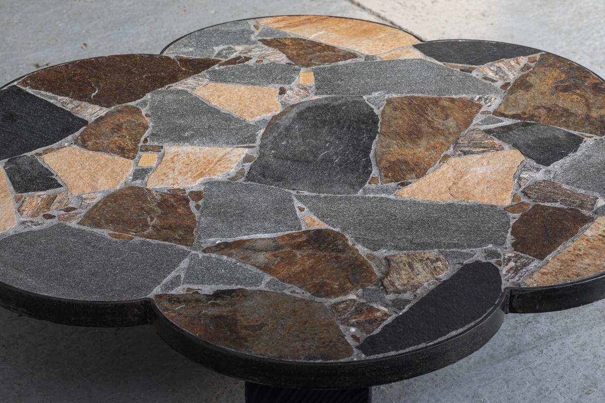 Brutalist slate stone coffee table designed and produced around 1950. A flower-shaped table top with an inlay of different kinds of stone, giving it a terrazzo look. The table top is supported by 3 black, wooden blocks with a beautiful hammered
