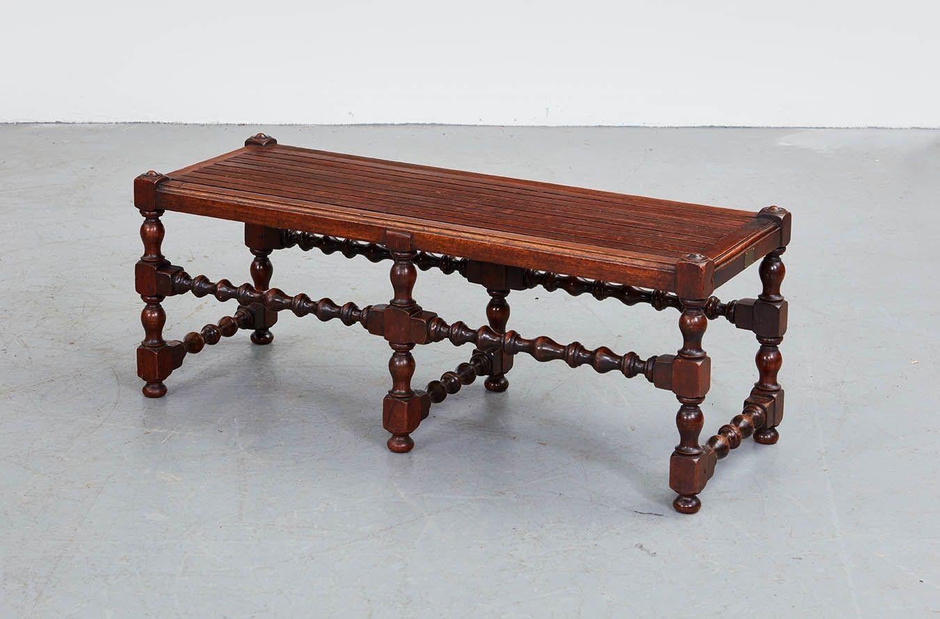 A long low luggage bench with flat slatted top over six bobbin legs joined by bobbin turned double side stretchers and central low cross bobbin stretcher. English, circa 1910. Bearing a brass maker's plate declaring: 