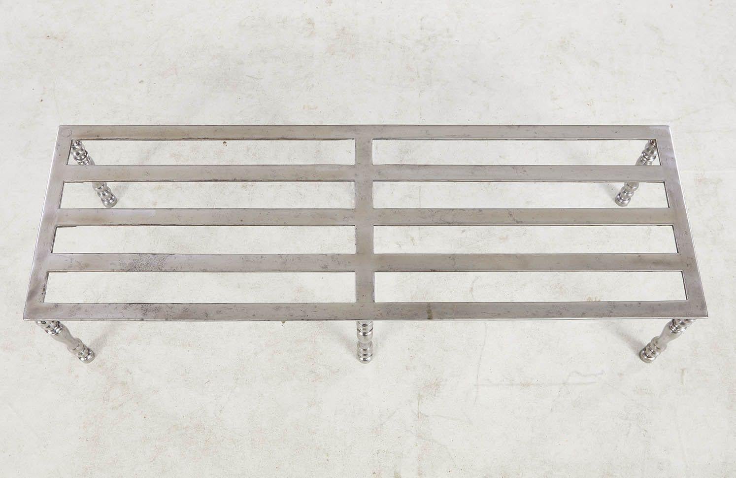 A long low stand having steel slats and borders with midpoint support on six turned steel legs, in a polished finish. Originally a grand fireplace trivet. Now useful as a luggage stand or a low drinks table.