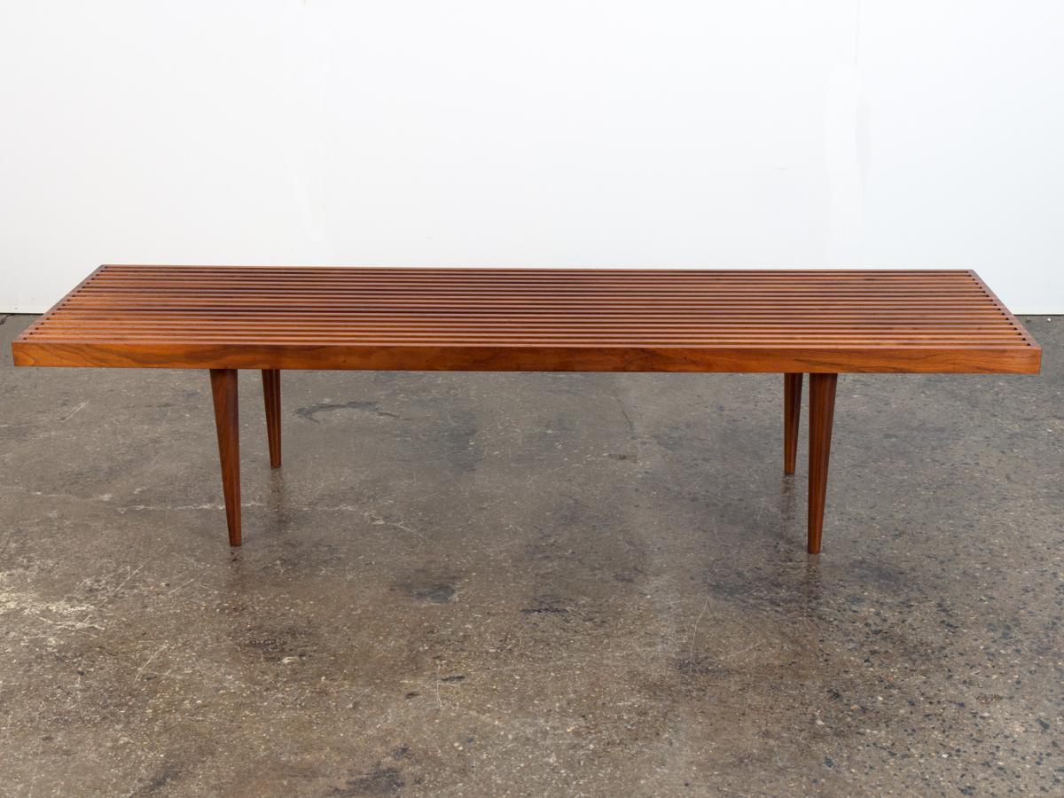Classic walnut slatted bench designed by Mel Smilow. Exceptionally crafted, with beautiful joinery, slender tapered legs and attractive wood grain throughout. A versatile piece that can be used as a coffee table. Wonderful condition, strongly built