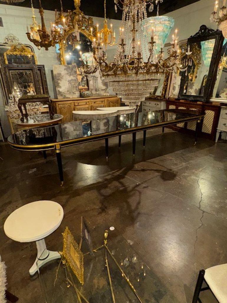 Esta elegante mesa de comedor de estilo Maison Jansen presenta un llamativo acabado lacado en negro piano con detalles de latón, que desprende un aspecto elegante y sofisticado. El diseño de la mesa recuerda a las piezas antiguas, añadiendo un toque