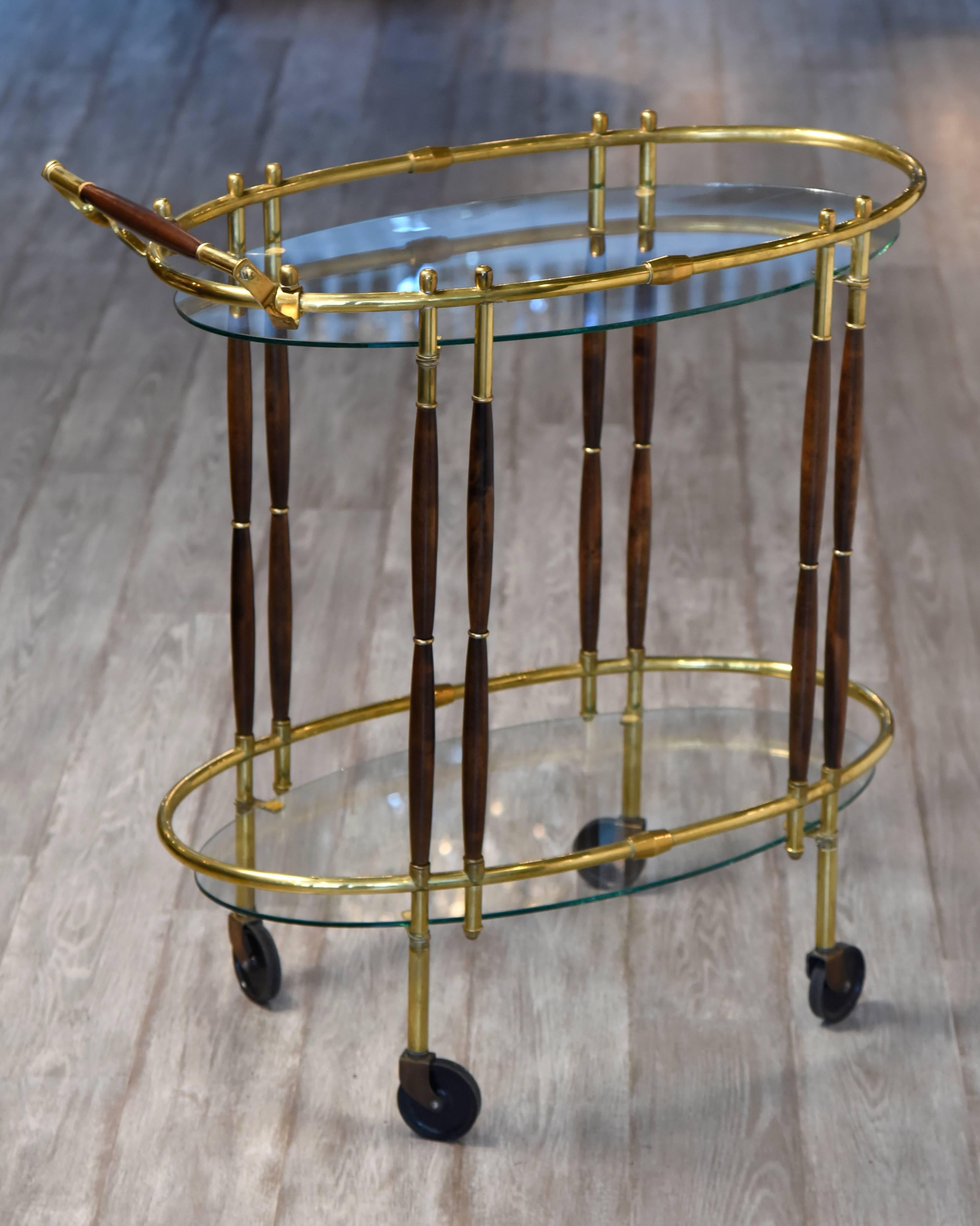 Sophisticated elegant oval bar cart having two glass tiers with brass galleries and sinuous walnut rods that link top to bottom. Measures: 33 inches high to handle. On wheels for easy moving.