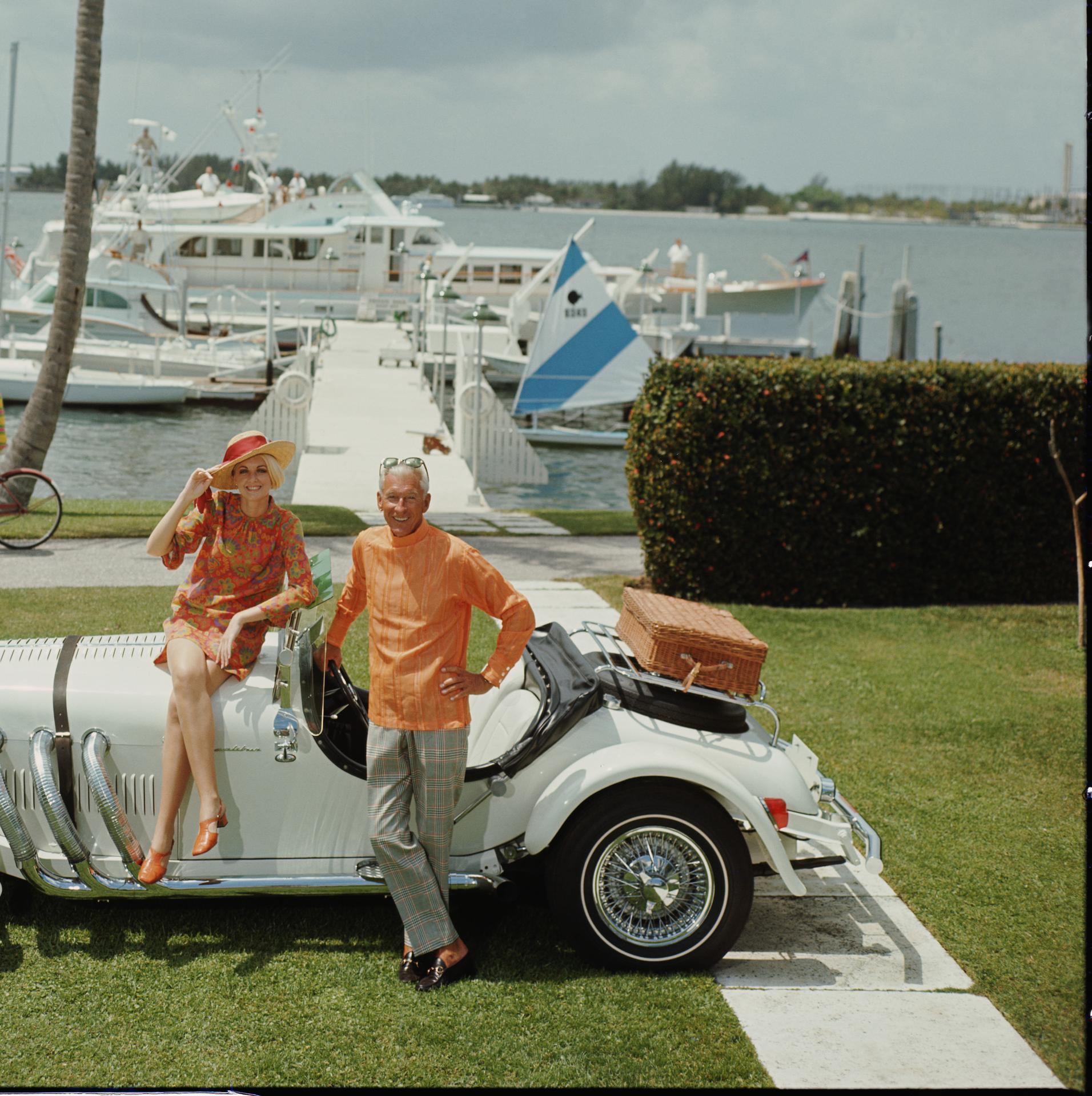 Questa fotografia figurativa della fine degli anni '60, scattata dal fotografo di società Slim Aarons, ritrae Jim Kimberly e sua moglie, con la sua auto sportiva bianca e le sue barche bianche ormeggiate sul lago Worth. È una socialite di Palm Beach
