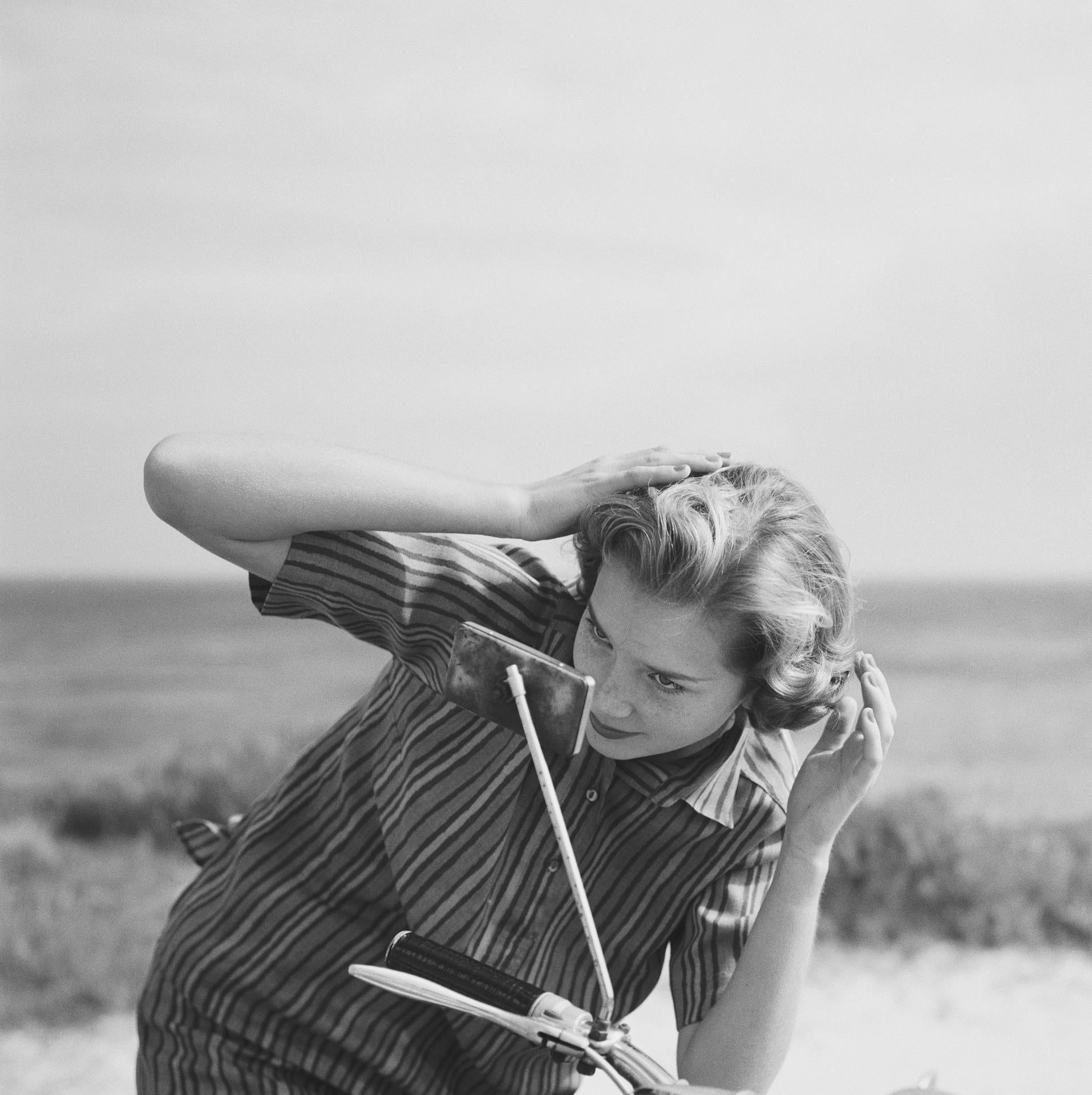 Esta fotografía de retrato urbano de finales de la década de 1950, captada por el fotógrafo de sociedad Slim Aarons, muestra a Faith Gibbons revisándose el pelo en el espejo de una moto, en las Bermudas. Lleva una sobrecamisa de lino irlandés a