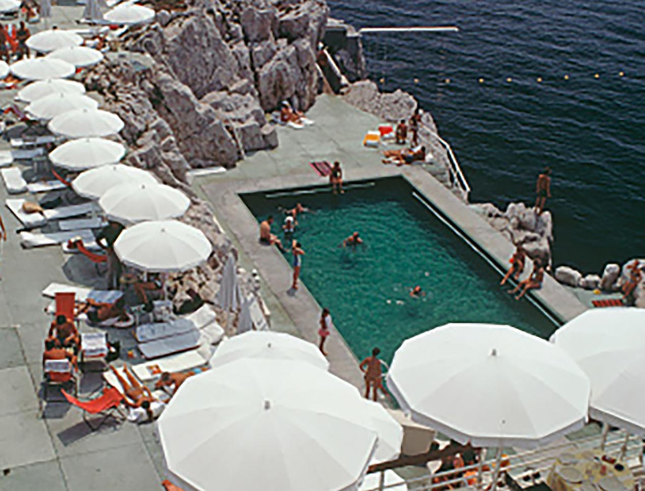 Cette photographie de paysage de la fin des années 1960, prise par le photographe mondain Slim Aarons, montre des clients autour de la piscine de l'hôtel du Cap Eden-Roc, à Antibes, en France, au mois d'août.

Il s'agit d'une édition de 150