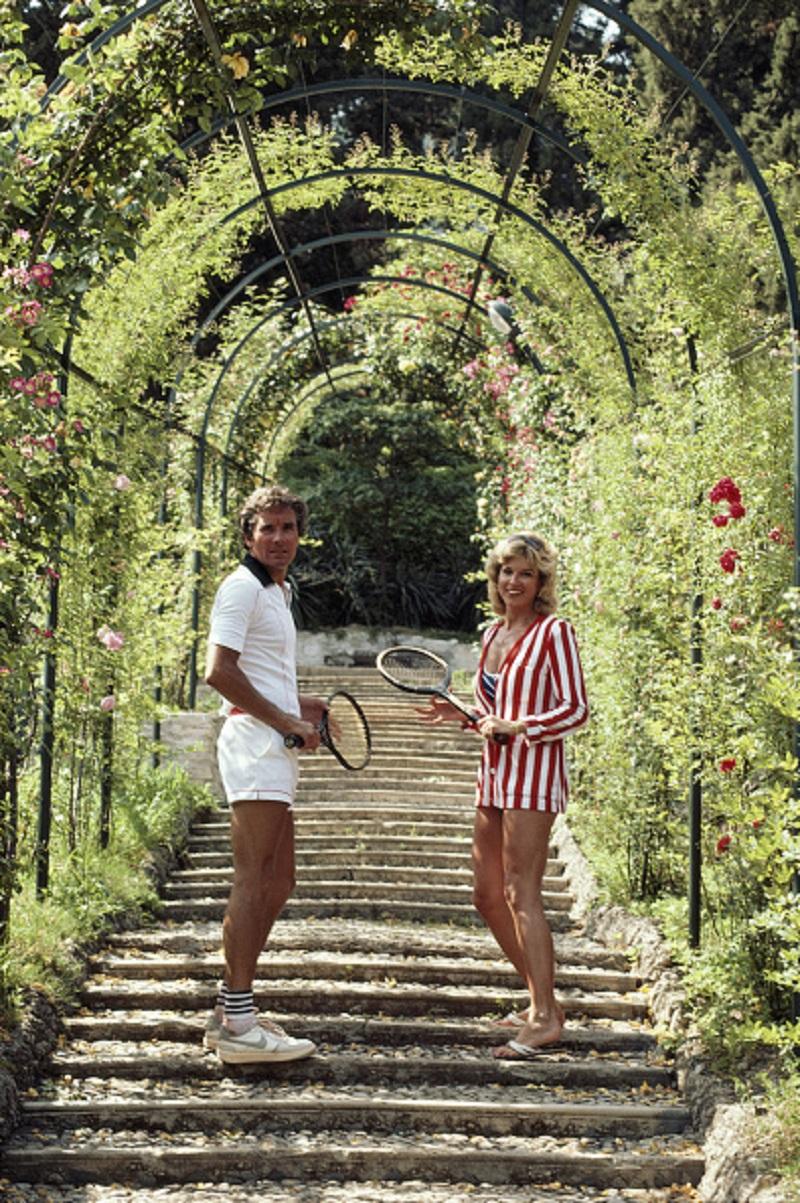 'Jack And GeeGee Entz' 1983 Slim Aarons Limited Estate Edition Print 

Jack and GeeGee Entz beneath the rose-canopied steps which lead to the entrance of the tennis courts at the Villa d'Este in Cernobbio, near Lake Como, Italy, circa