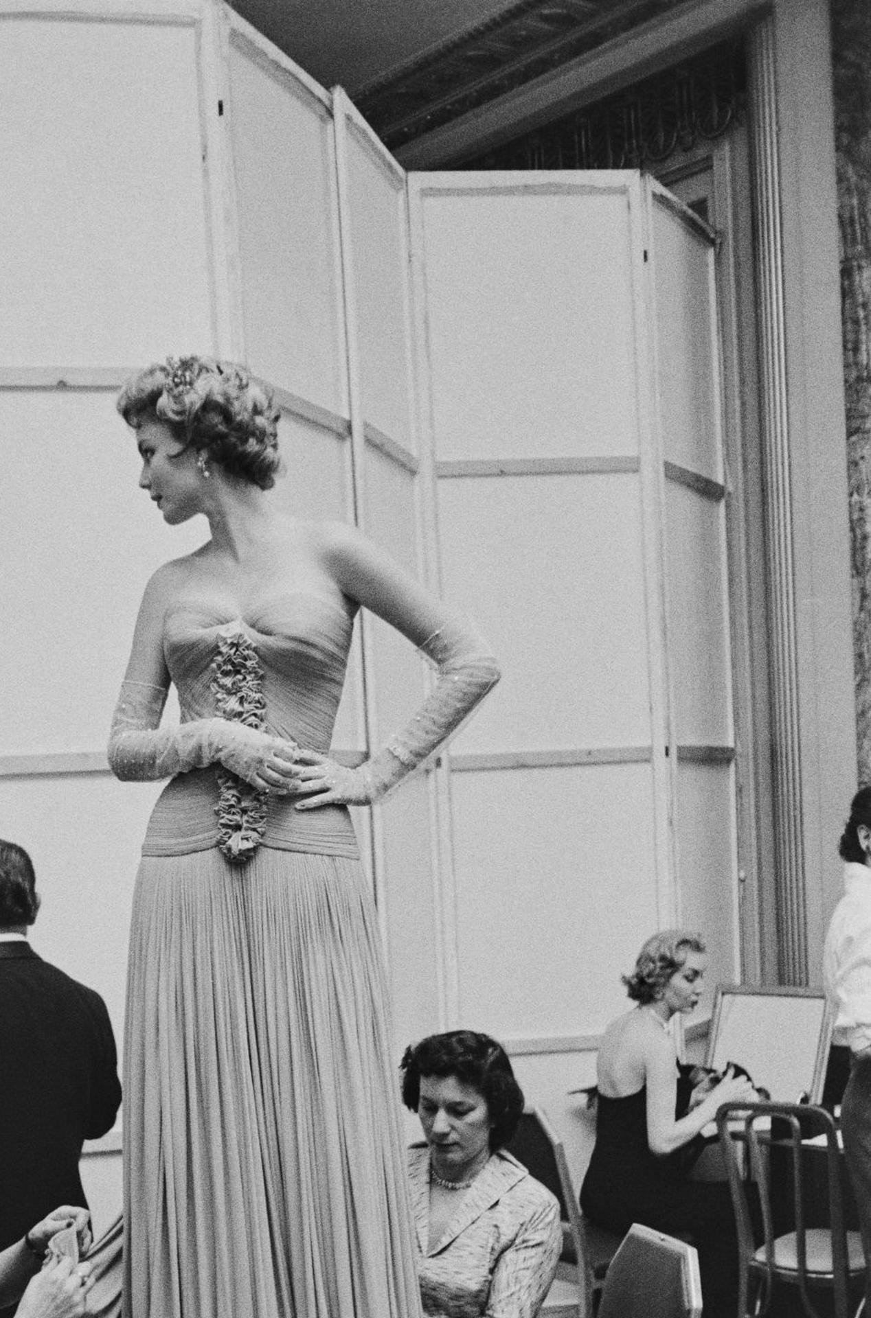 Dieses Porträtfoto des Society-Fotografen Slim Aarons aus der Mitte der 1950er Jahre zeigt eine junge Frau, die ihr Kleid für einen Debütantenball in letzter Minute ändern lässt.

Dies ist eine nachlassgestempelte und handnummerierte Auflage von 150