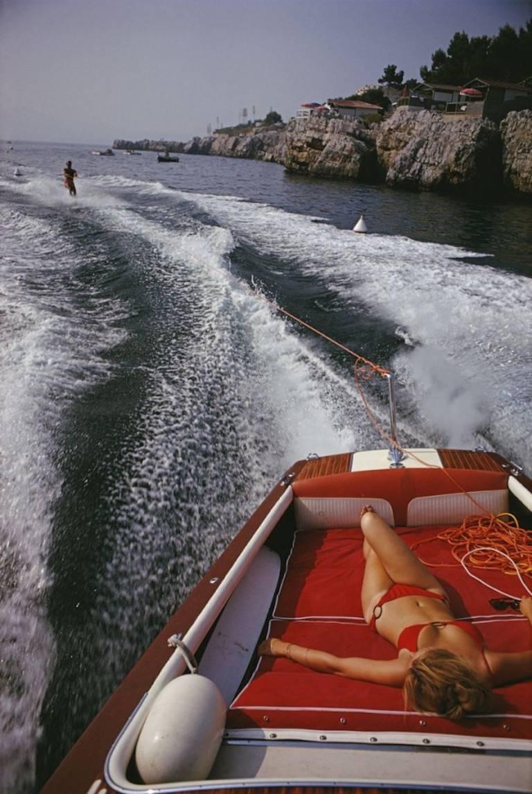 freizeit in Antibes" (Slim Aarons Estate Edition)

Eine Frau sonnt sich in einem Motorboot, das einen Wasserskifahrer zieht, auf dem Meer vor dem Hotel du Cap-Eden-Roc in Antibes an der Côte d'Azur, August 1969.

Glamouröser und dekadenter geht es