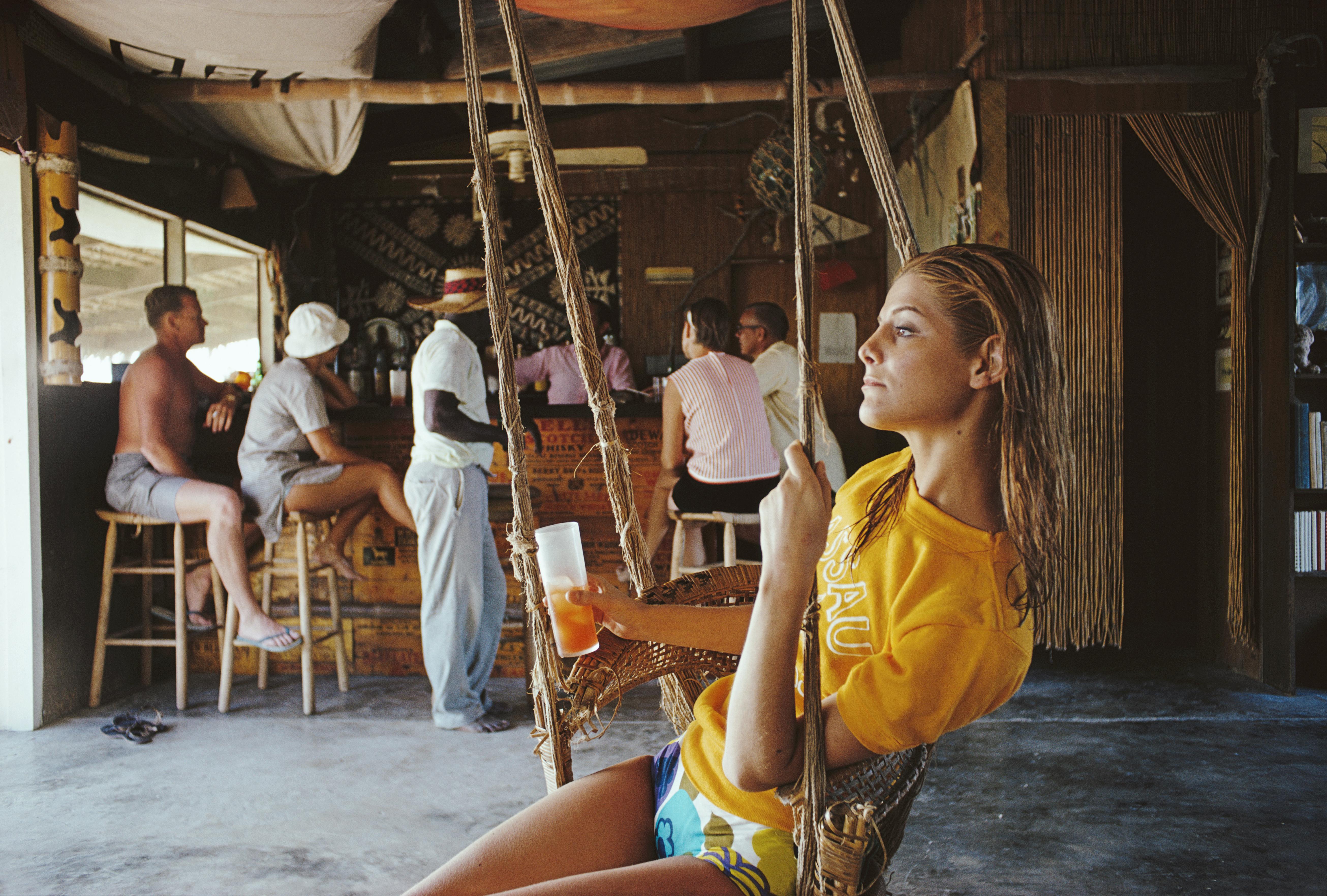 'Life In The Bahamas' 1967 Slim Aarons Limited Estate Edition Print 

A woman sitting in a hanging chair while having a drink in a bar in the Bahamas, in April 1967. A group of drinkers stand and sit at the bar in the background. 

Produced from the