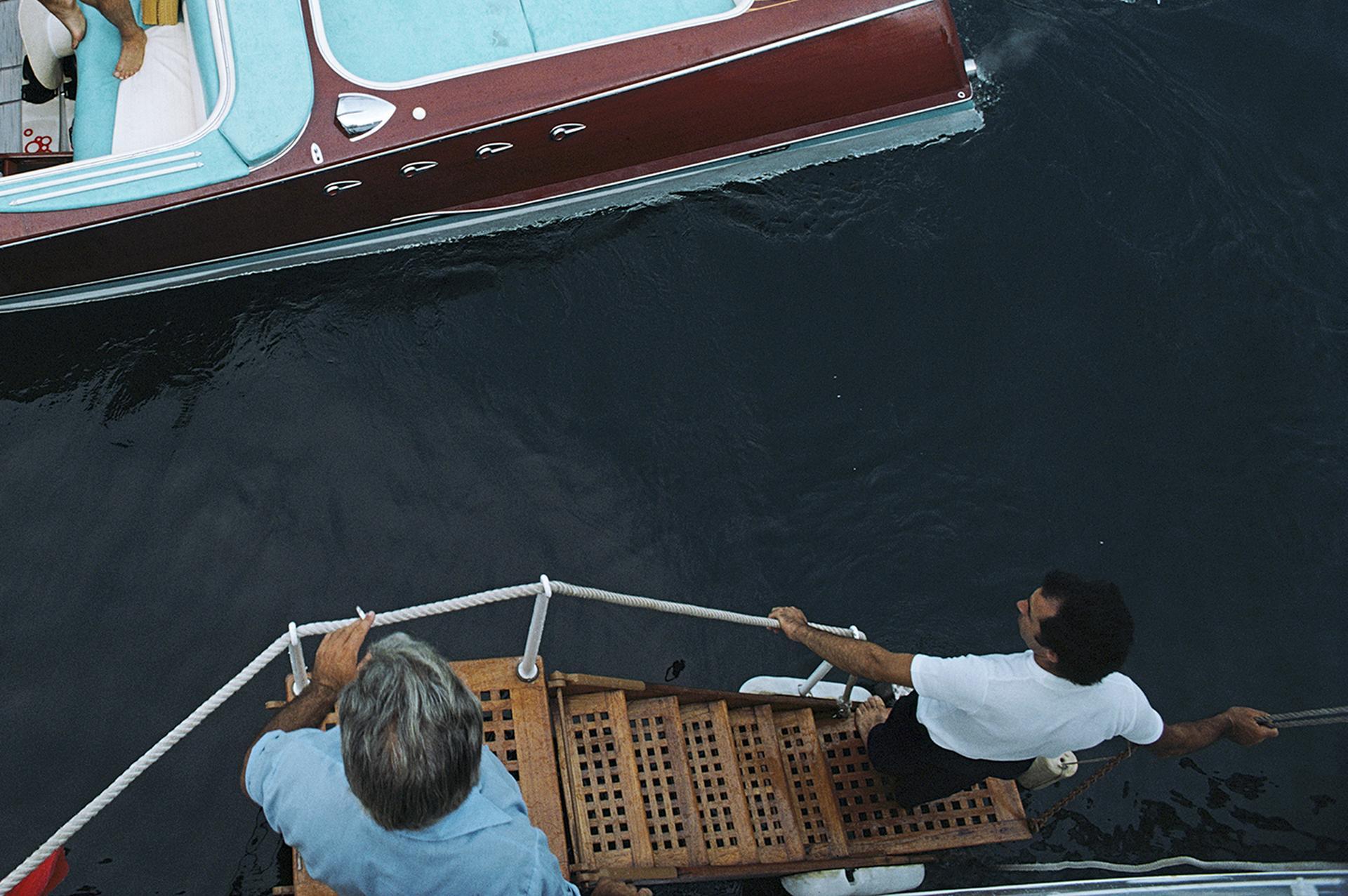Weiß gekleidete Freunde besteigen ein Riva-Boot in Monte Carlo, Monaco, 1975.

In dieser klassischen Slim-Aarons-Szene im Glamour der 1970er Jahre setzen weiße Vintage-Mode und türkisfarbene Polstermöbel farbliche Akzente zum polierten Holz und den
