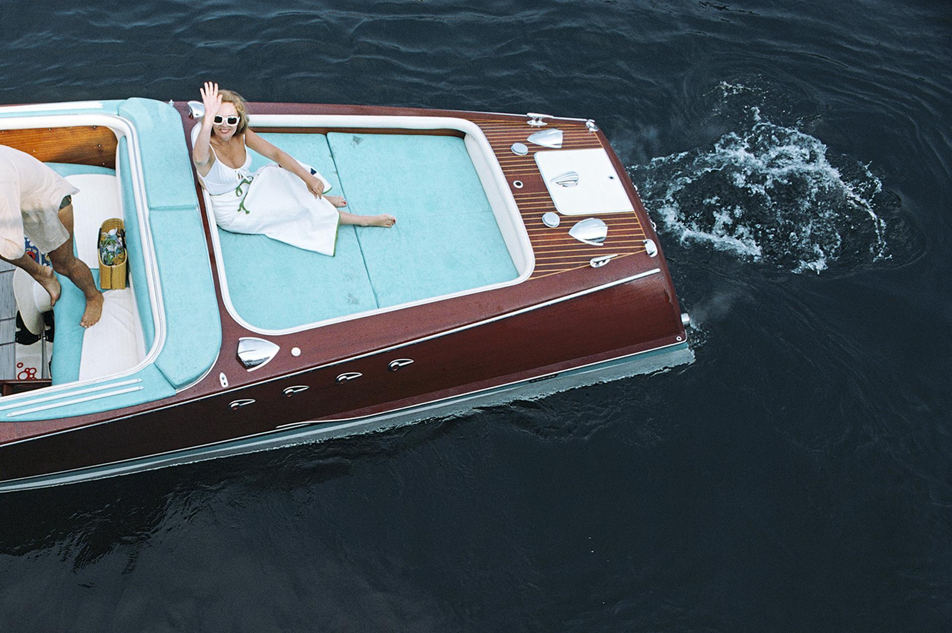 Weiß gekleidete Freunde entern ein Riva-Boot in Monte Carlo, Monaco, 1975.

In dieser klassischen Slim-Aarons-Szene des Glamours der 1970er Jahre setzen weiße Vintage-Mode und türkisfarbene Polstermöbel farbliche Akzente zum polierten Holz und den