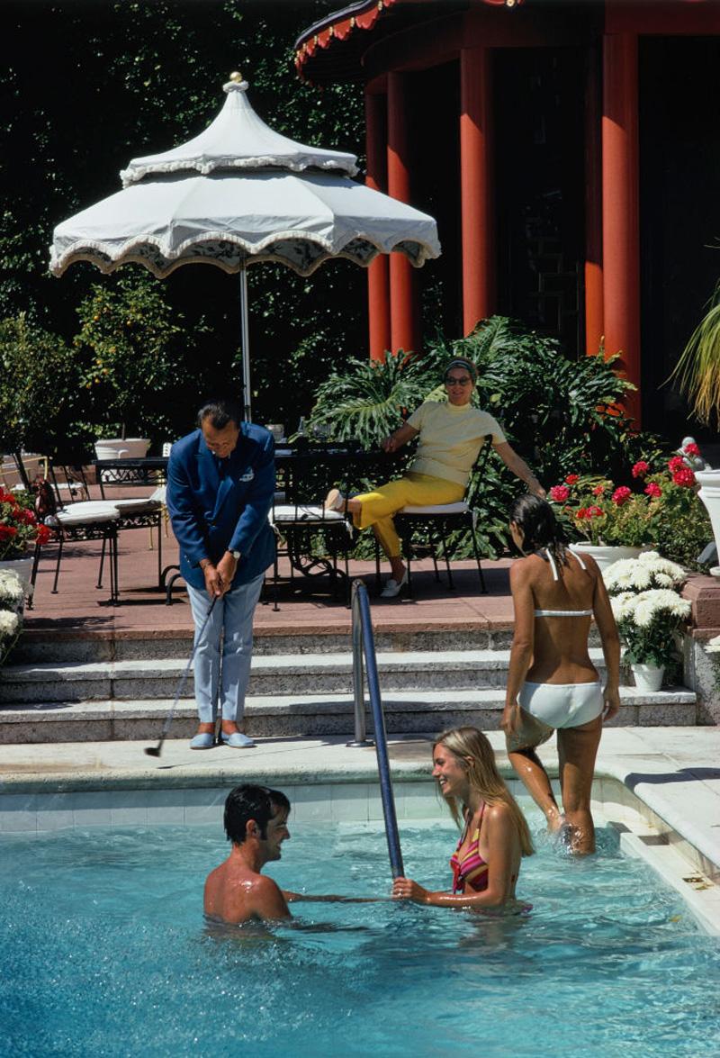Ein Mann hält einen Golfschläger am Pool, während der amerikanische Schauspieler Lee Kinsolving mit seiner Lebensgefährtin Lillian Kinsolving (geborene Crawford) am Pool des Hauses von Albin und Margo Lykes Holder in Palm Beach, Florida, im Mai 1970
