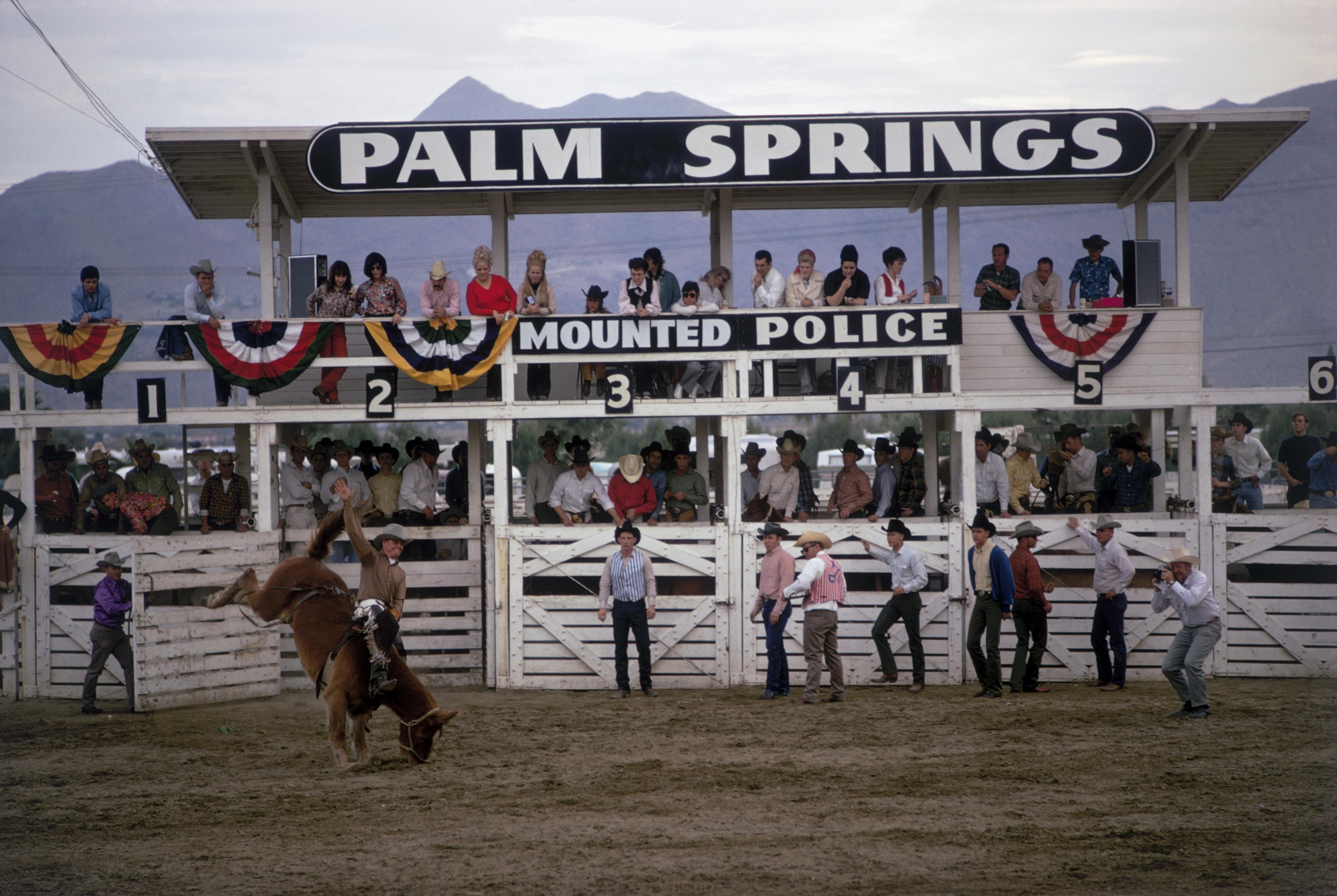 palm Springs Rodeo' 1970 Slim Aarons Limitierte Nachlassausgabe Druck 

Zuschauer beobachten von der Tribüne aus, wie ein Teilnehmer beim Palm Springs Mounted Police Rodeo, Palm Springs, Kalifornien, Januar 1970, ein bockendes Pferd reitet. 
3(Foto: