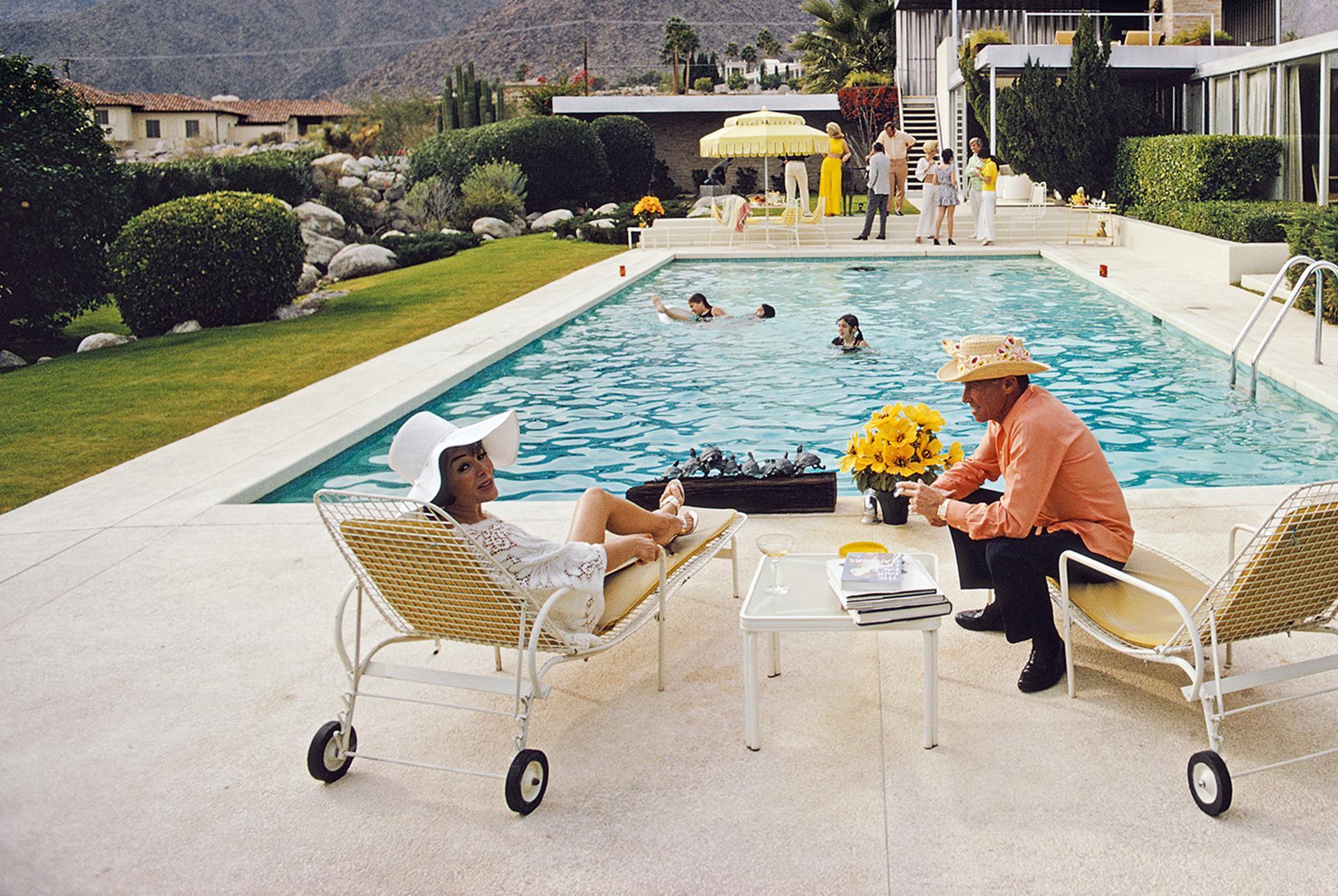 This early 1970s lifestyle photograph, captured by society photographer Slim Aarons, features Lita Baron talking with a guest at a poolside party at Nelda Linsk's desert house in Palm Springs, California, January 1970. The house was designed by