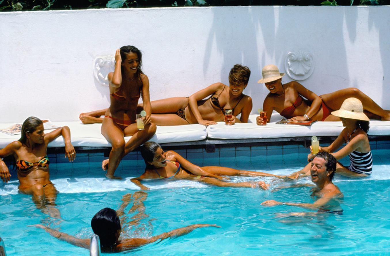 Pousada Dos Buzios 
1975
by Slim Aarons

Slim Aarons Limited Estate Edition

Friends by the pool at Pousada Dos Buzios in Brazil, August 1975. Left is student Fabian Crespo, lying on the cushions (without the hat) is Tania Louise, the man with just