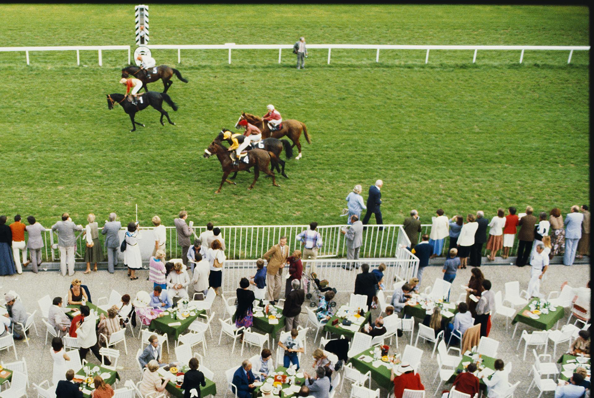 Cette photographie de paysage de la fin des années 1970, prise par le photographe mondain Slim Aarons, montre des spectateurs dînant en plein air pendant que les chevaux passent à l'hippodrome de Baden Baden, en Allemagne.

Il s'agit d'une édition