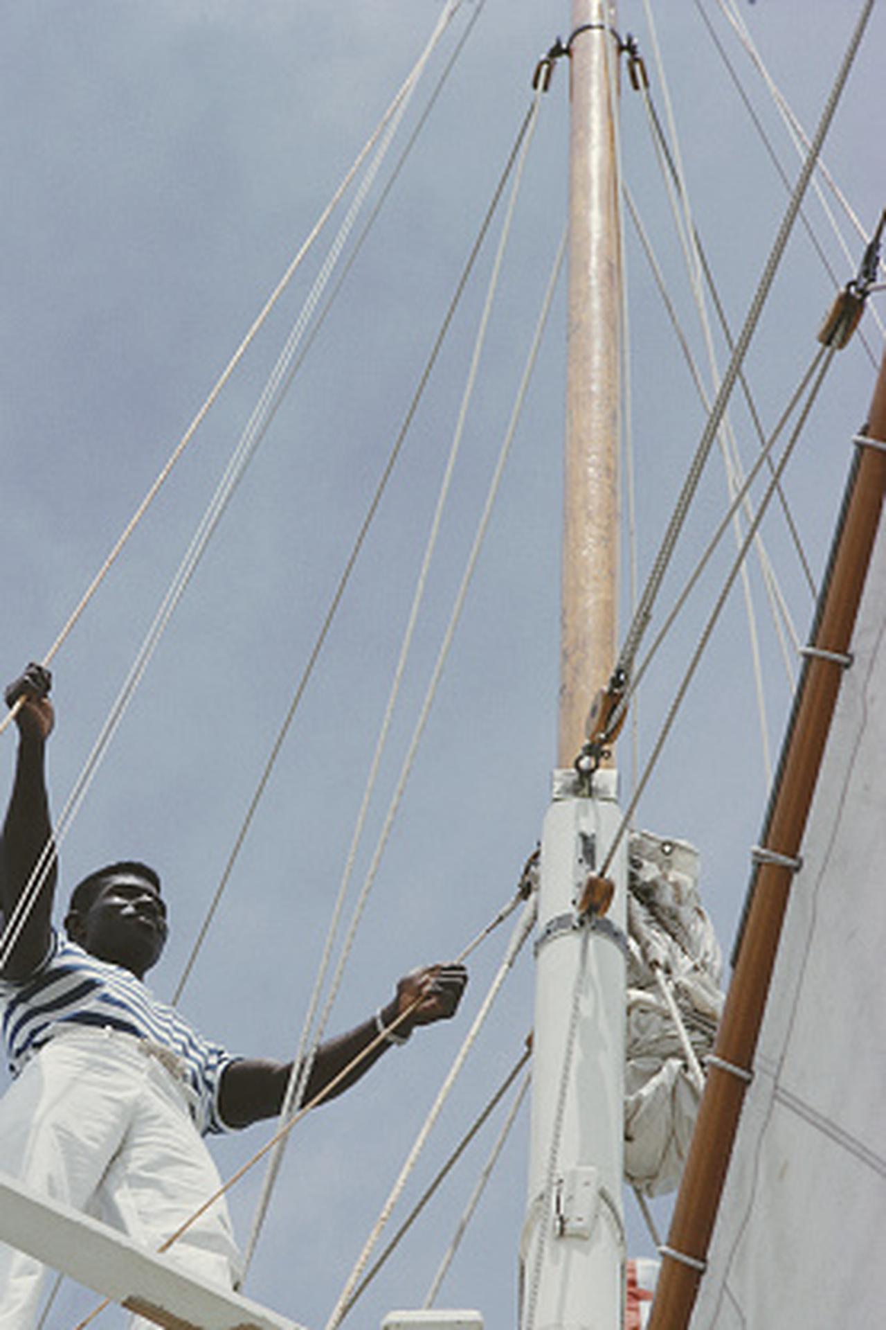 Dieses Porträtfoto des Gesellschaftsfotografen Slim Aarons aus den späten 1960er Jahren zeigt ein Besatzungsmitglied, das sich um die Takelage einer Yacht in Lyford Cay auf den Bahamas kümmert.

Dies ist eine nachlassgestempelte und handnummerierte