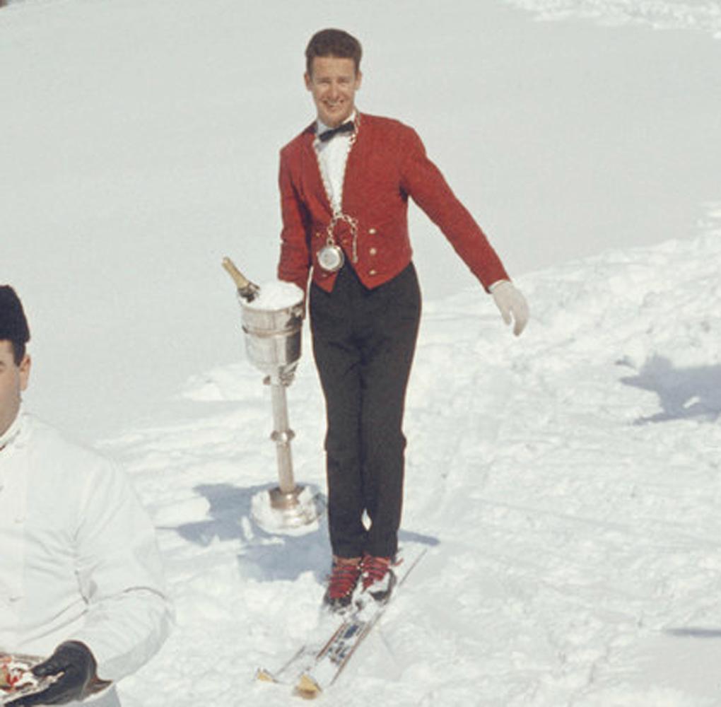 Trois serveurs de ski sur une piste de ski, vers 1960. Au premier plan, un chef vêtu de blanc se dirige vers le spectateur en skiant, offrant un oiseau magnifiquement préparé sur un plateau. Derrière lui, un homme en veste rouge bordeaux formelle