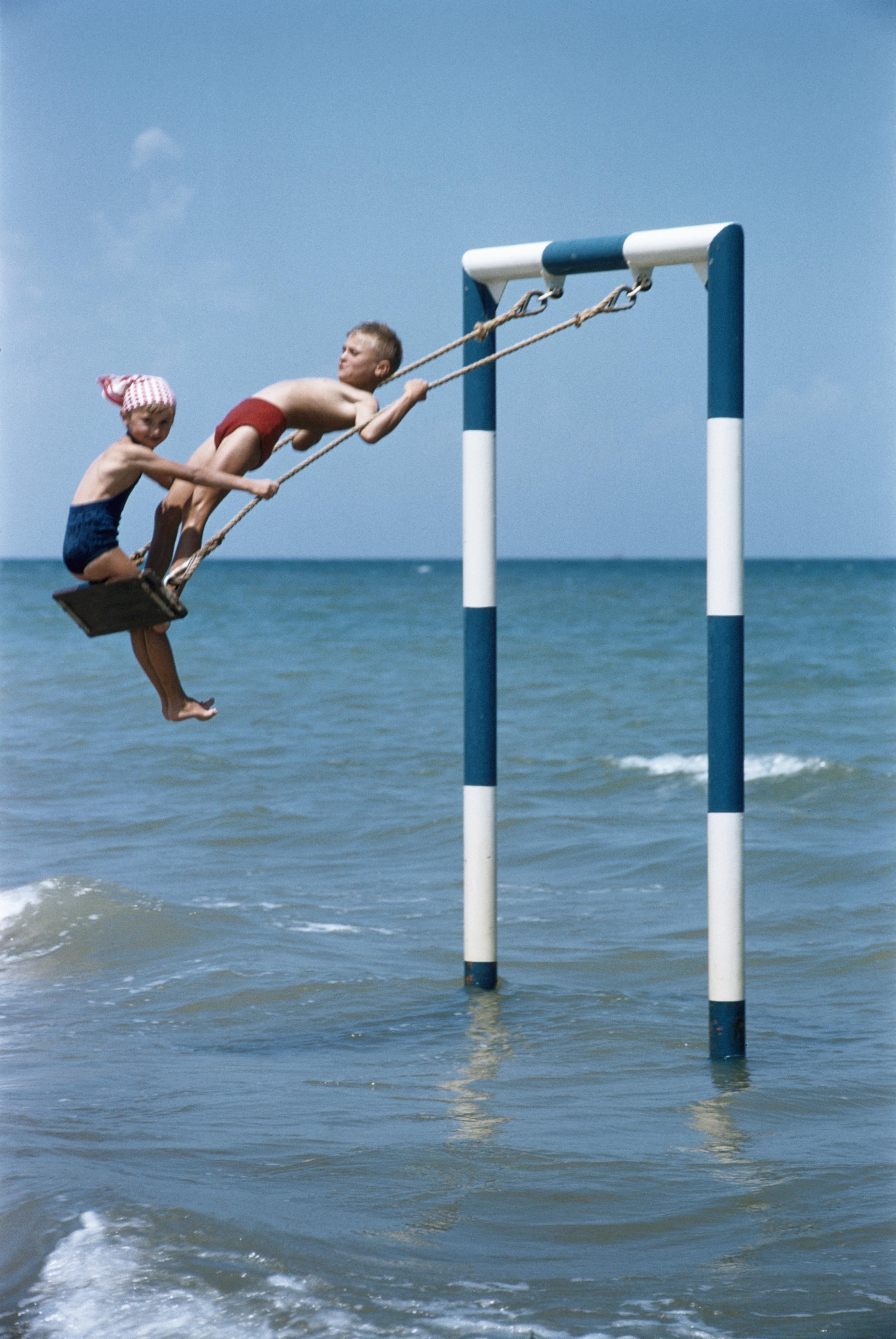 Slim Aarons
Adriatic Riviera
1956
Chromogenic Lambda Print
Estate stamped and hand numbered edition of 150 with certificate of authenticity from the estate. 

Two children playing on a swing in the waters on the coast of Rivabella, near Rimini, on