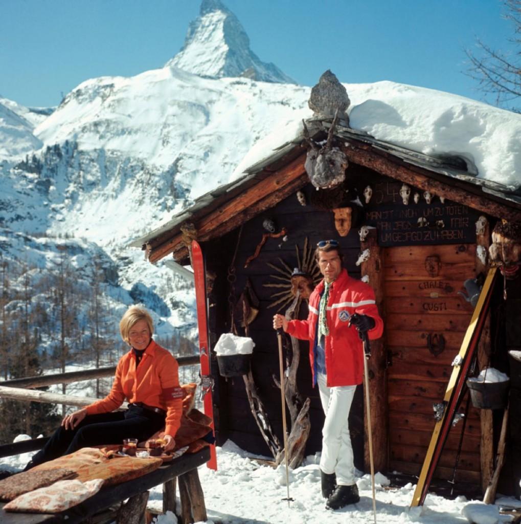 'Chalet Costi' by Slim Aarons

Skiers outside the Chalet Costi in Zermatt, 1968.

Stylishly dressed skiers take a break from the slopes outside a wooden cabin with the snow covered mountains visible in the background behind them.

Typically 'Slim'
