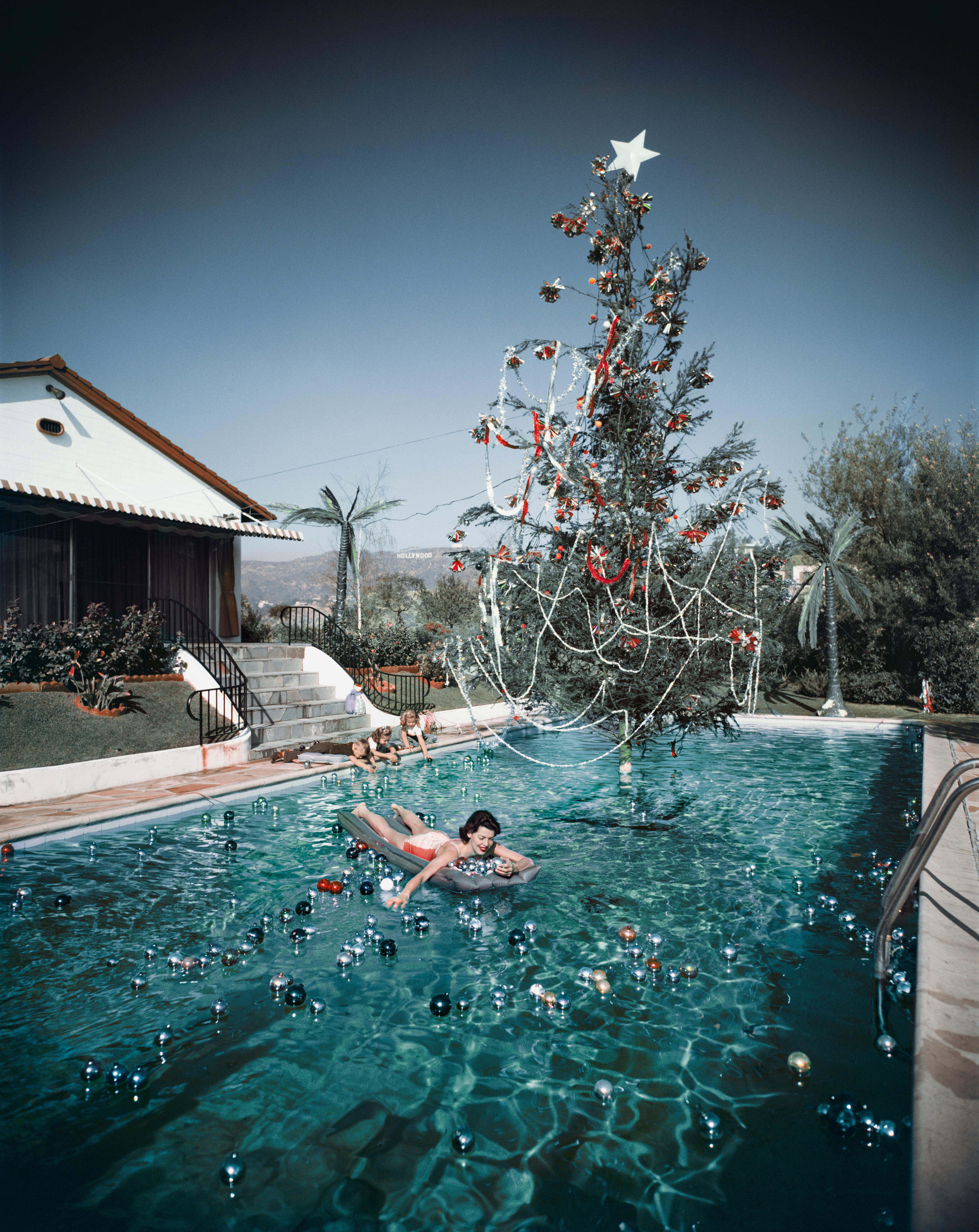 Rita Aarons, Ehefrau des Fotografen Slim Aarons, auf einer Luftmatratze in einem weihnachtlich geschmückten Swimmingpool, Hollywood, 1954. In der Ferne ist das Hollywood-Schild zu sehen. 

Nachlassgestempelte und handnummerierte Auflage von 150