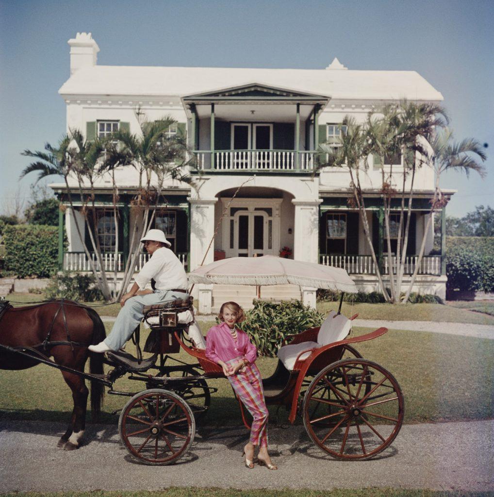 Bermudanische Hostess 

Polly Trott Hornburg vor dem typischen Haus ihres Vaters auf den Bermudas. Sie trägt eine von ihr selbst entworfene Thaibok-Hose und ein Hemd für eine Fahrt in einer offenen Kutsche, Bermuda, 1957.

Wunderschöner Druck im