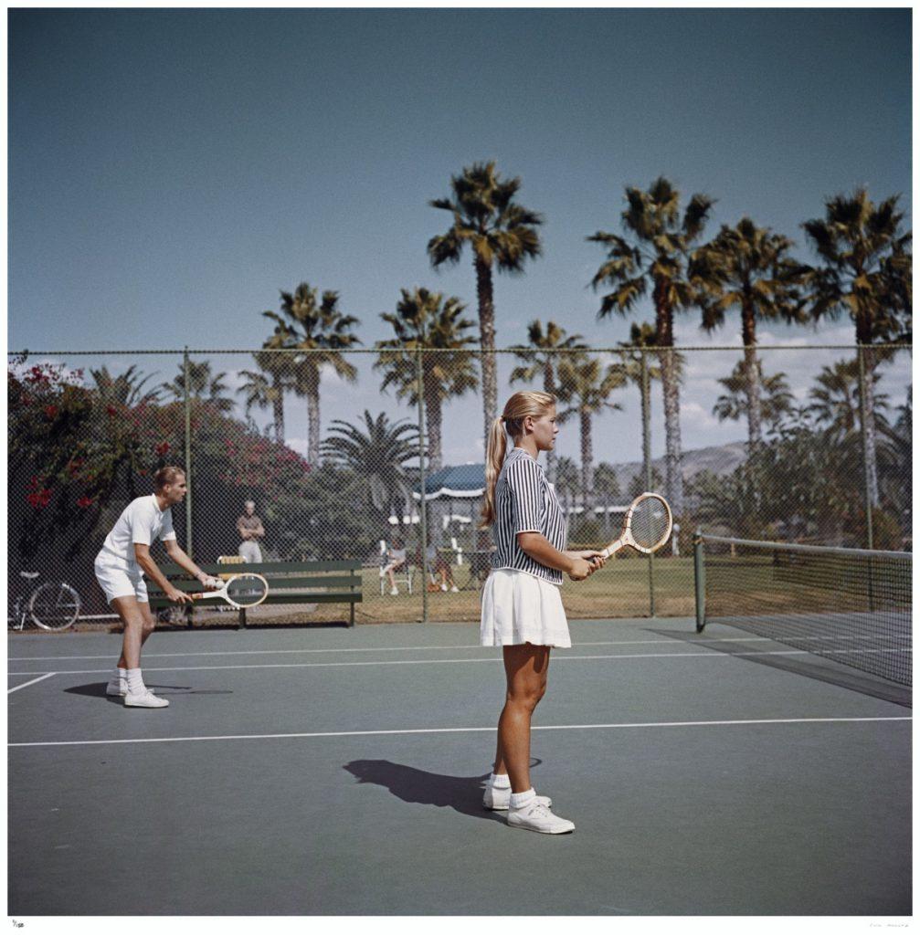 Slim Aarons Estate Edition -  Taking The Plunge 

Limited Edition Estate Stamped Print (edition size 1/150).

A man and a woman playing tennis on a court surrounded by palm trees, San Diego, California, October 1956.

This photograph epitomises the