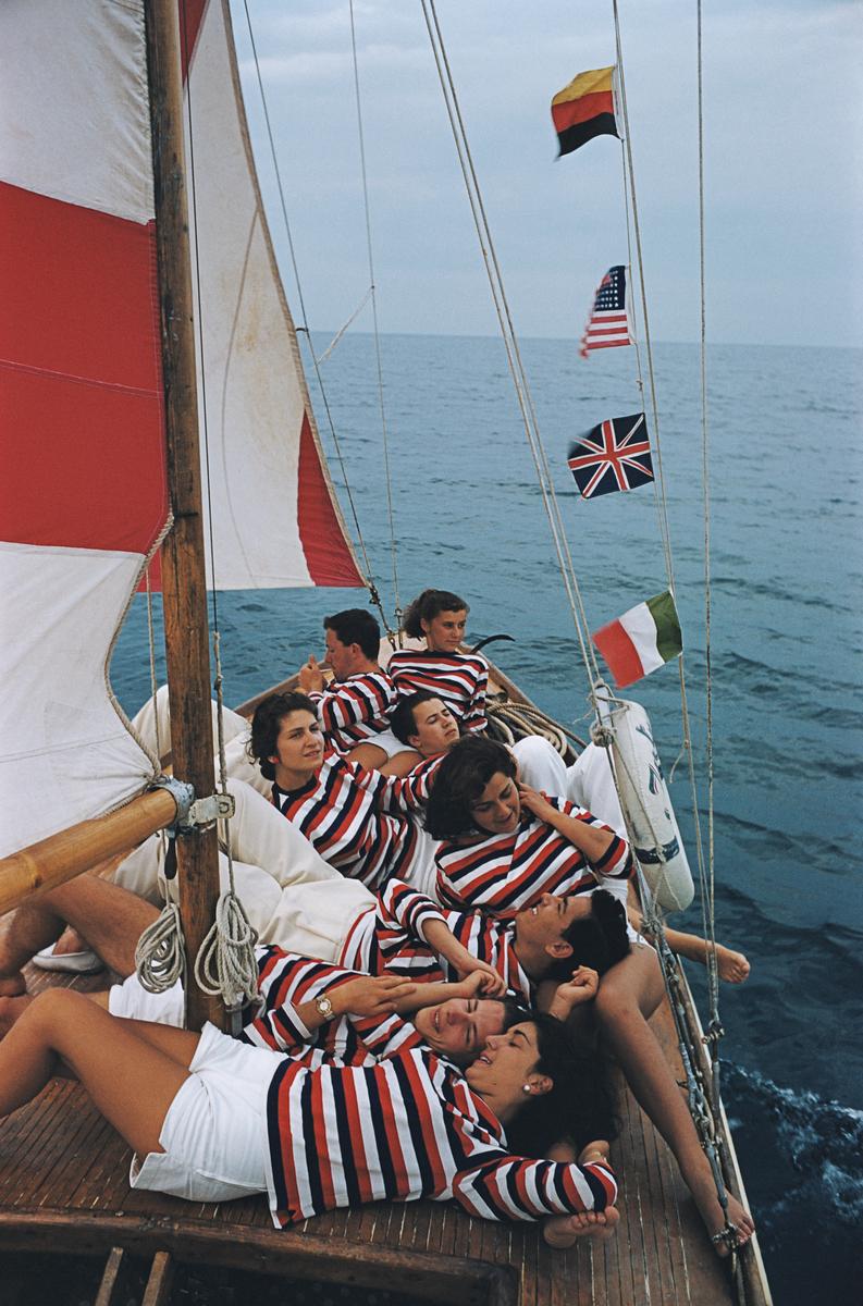 Riviera Adriática

Hombres y mujeres -vestidos con camisetas a rayas rojas, blancas y azules- descansando en un pequeño barco, con un mástil rojo y blanco, y pequeñas banderas de Alemania, EEUU, Gran Bretaña e Italia atadas a las jarcias, en la